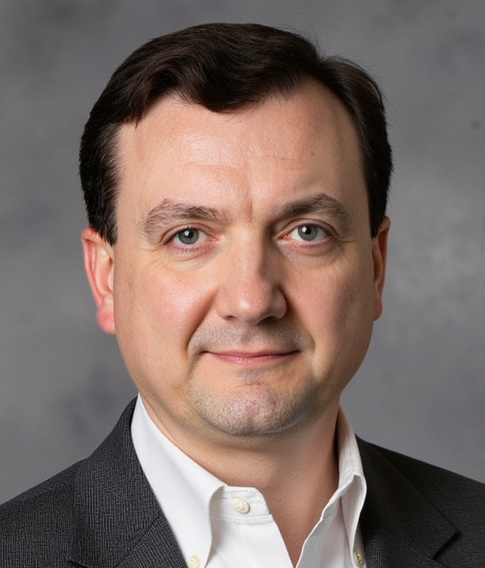 A headshot of a man with brown hair and green eyes. He is wearing a white dress shirt and a dark suit jacket. The background is a neutral gray.