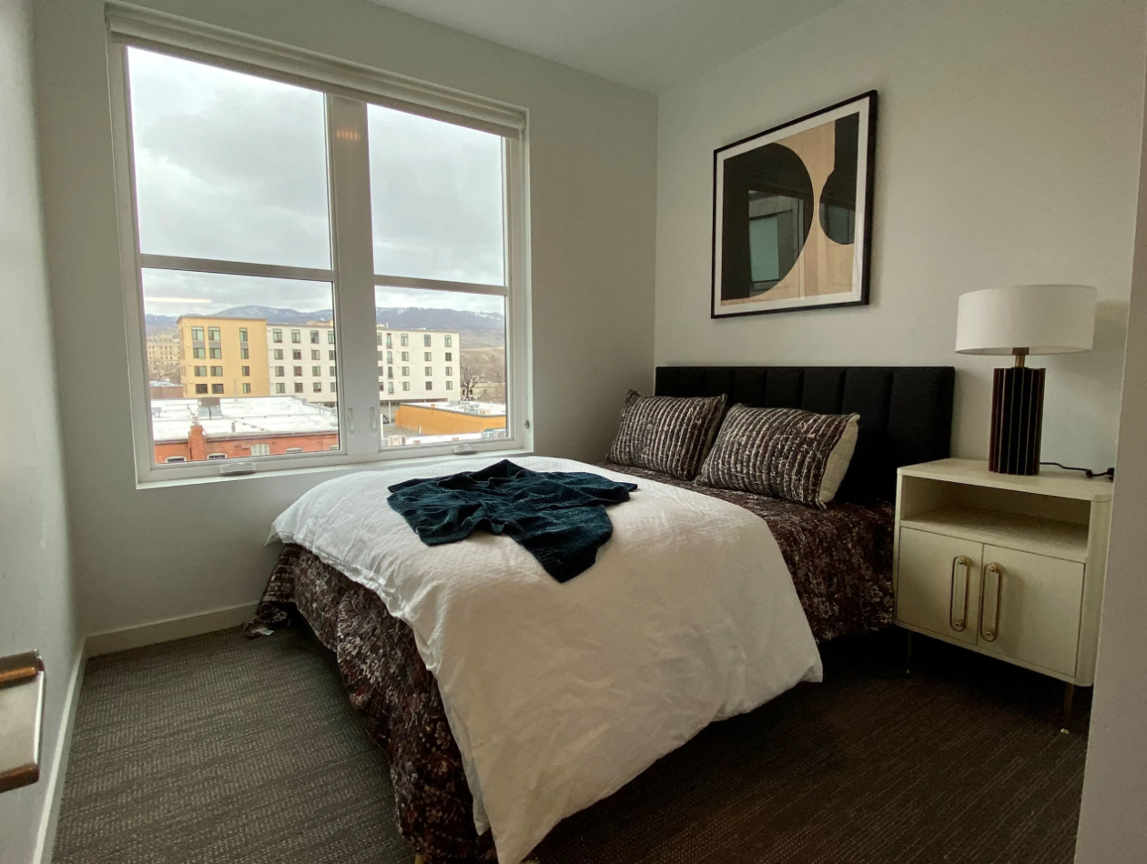 A cozy bedroom with oversized windows that view the foothills of Boise with a bed, mightsand with gold hardware, and desk lamp