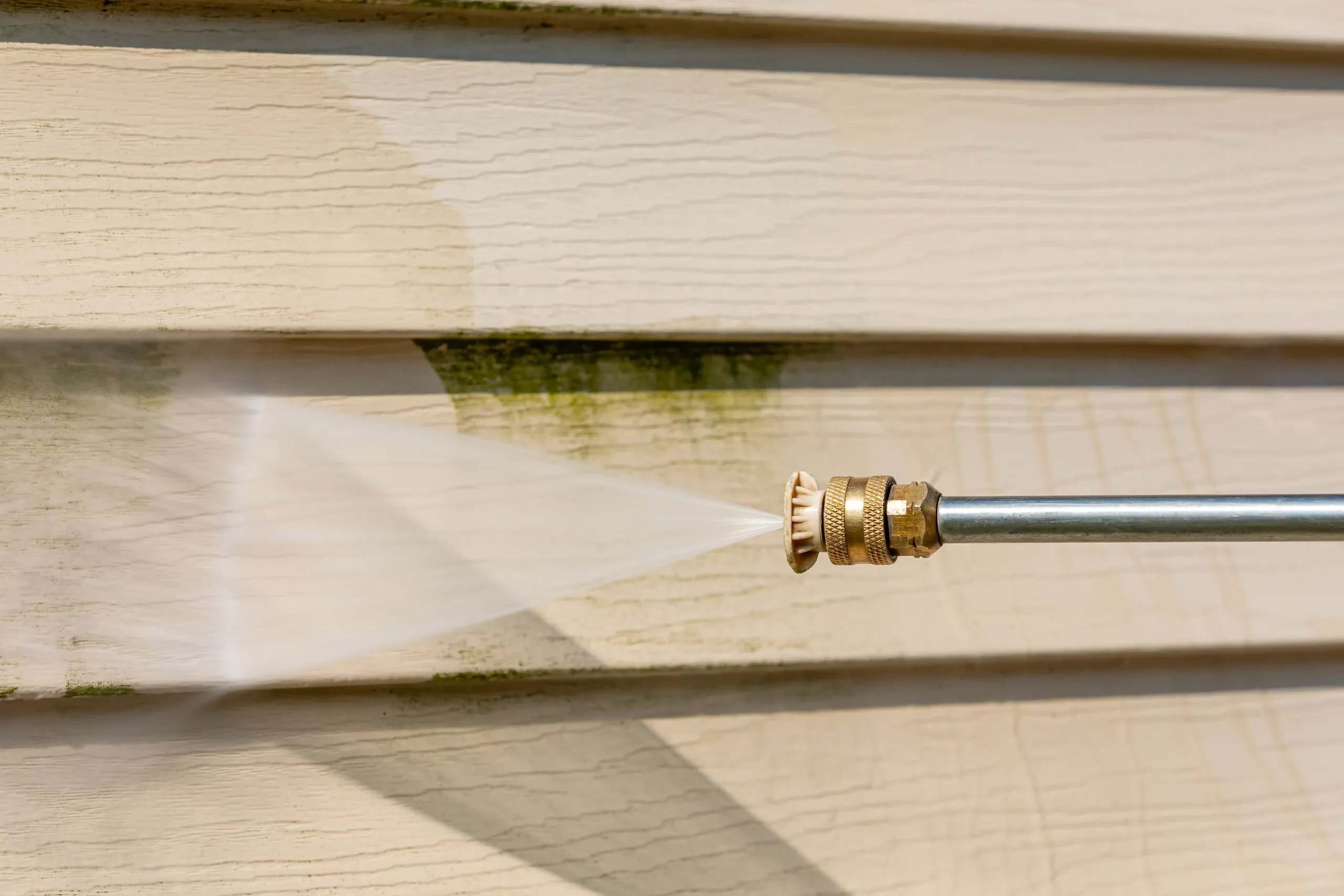 A close-up shot shows a pressure washer spray cleaning a beige vinyl siding of a house, removing green algae and dirt from the surface. The water spray is focused and powerful, creating a mist and revealing a clean section of siding behind the dirty area. The siding has a wood-grain texture.