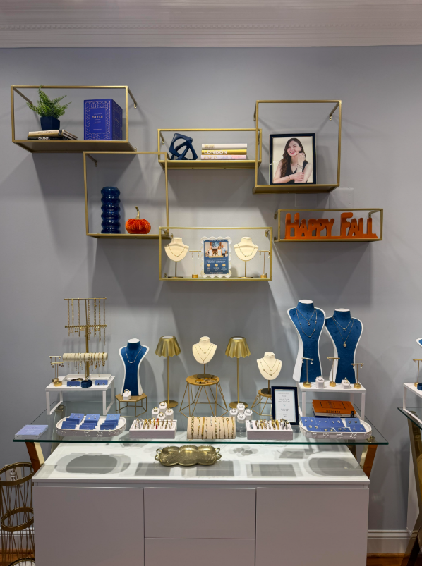Eliana Curated showcases necklaces, bracelets and earrings on blue and cream stands. Gold wall shelves decorative books and autumn accents create an inviting setting to explore unique jewelry designs.