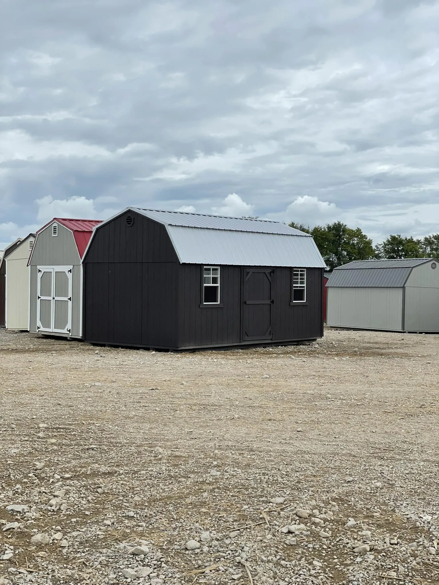 Black barn-style portable storage building with silver metal roof, centered entry door, and side windows. Positioned on gravel lot, offering durable weather-resistant storage with classic barn-inspired design.