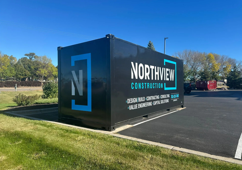 A black Northview Construction branded jobsite storage container positioned outdoors on asphalt next to a grassy lawn under a clear blue sky. The container displays the company logo, name, phone number, and listed services including Design/Build, Contracting, Consulting, Value Engineering, and Capital Solutions.