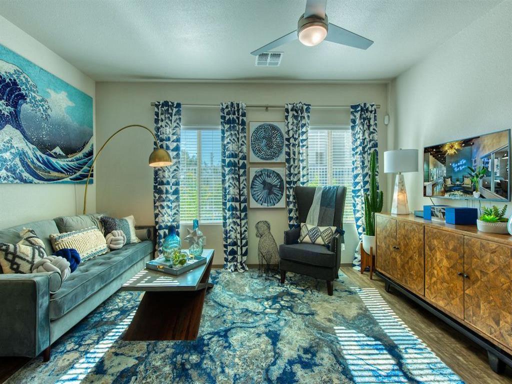 A living area of an apartment with wood floors with a blue patterned rug and a pair of windows with blue patterned curtains