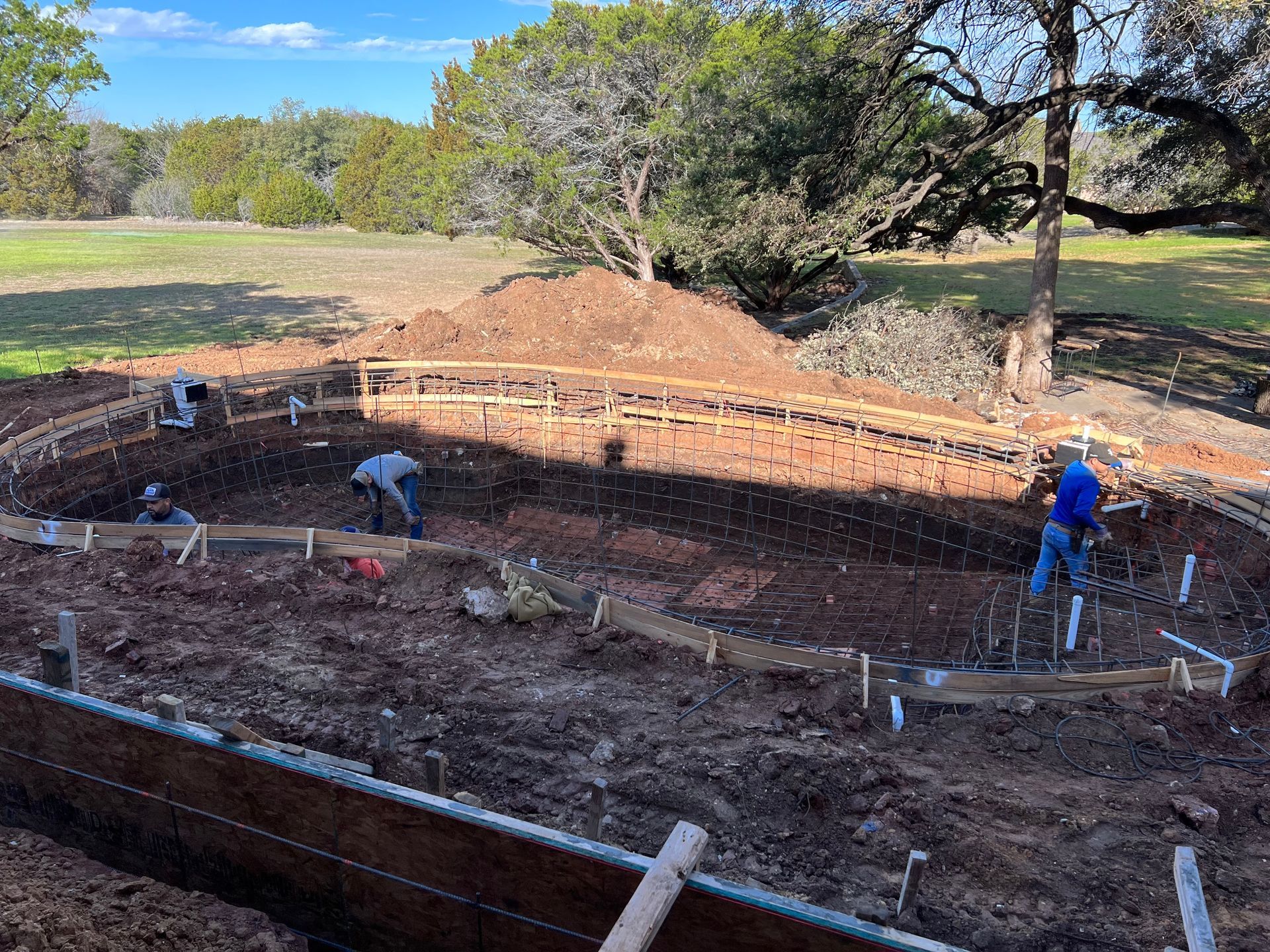 Construction workers install a dense steel rebar cage within a excavated pool site. The reinforced structure defines the pool's curved shape and depth before the concrete gunite is applied.