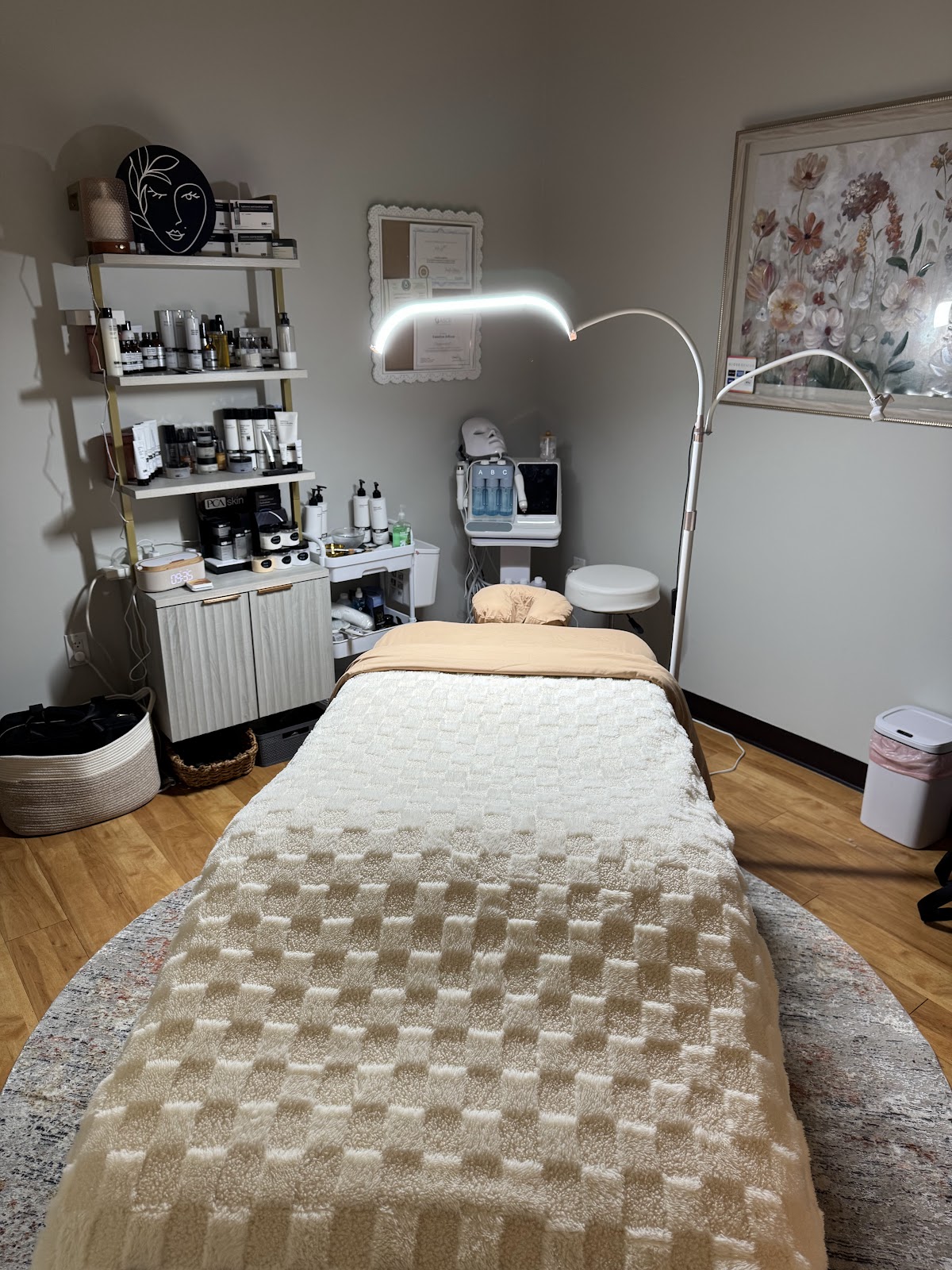 A serene spa room with a massage bed covered in a textured beige blanket, a shelf filled with skincare products, and a facial steamer with an LED lamp. A framed floral artwork hangs on the wall.