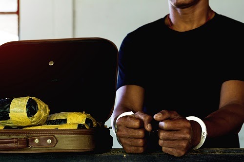 Handcuffed individual in a dark shirt beside an open suitcase with wrapped packages.