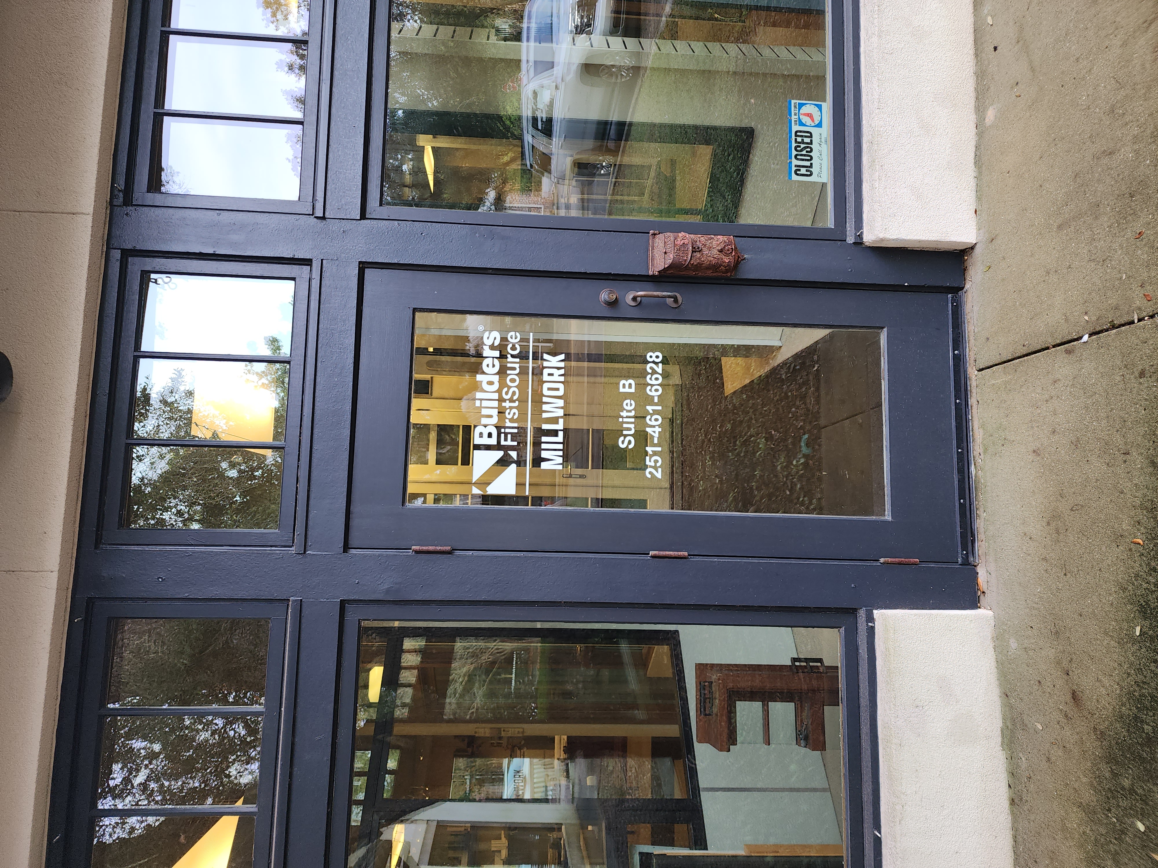 The front entrance of the Builders FirstSource Millwork office, labeled Suite B with the phone number 251-461-6628. The door features a clean, modern design with an open/closed sign displayed at the bottom, and reflections of the surrounding trees visible in the glass.