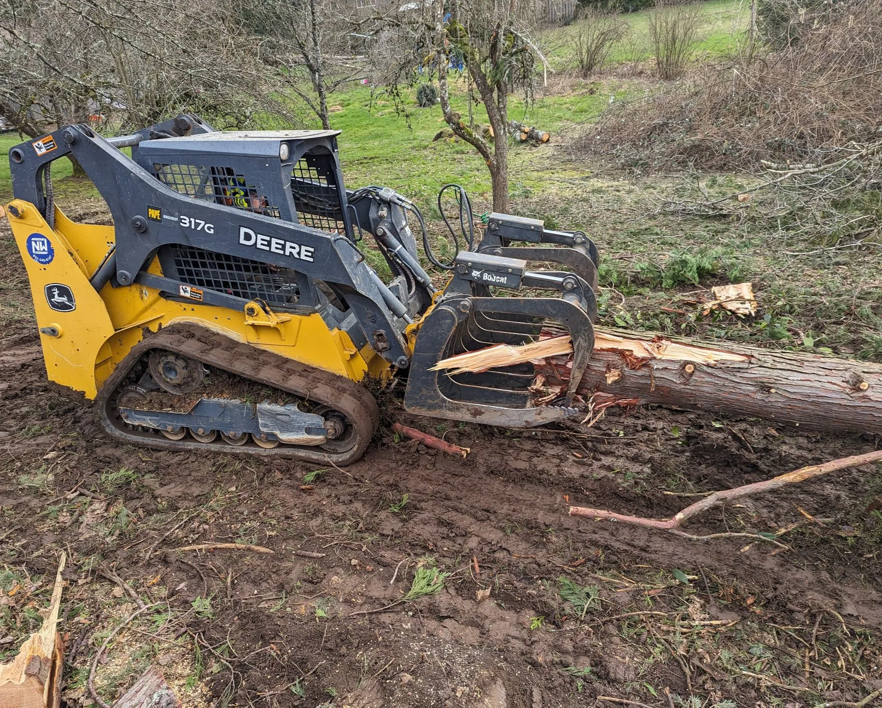 A tracked skid steer equipped with a grapple attachment moves a damaged tree trunk across muddy grou 