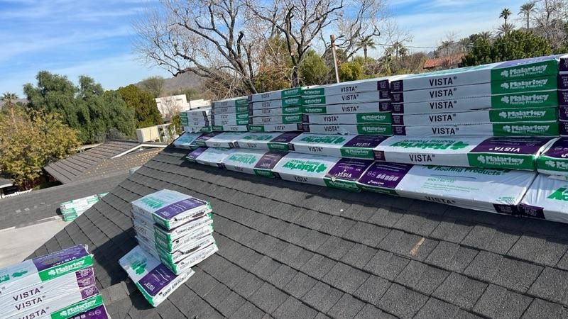 A high-angle rooftop view shows a professional roofing project in progress. Multiple stacks of Malarkey Vista asphalt shingle bundles are neatly staged along the roof ridge against a clear blue sky.