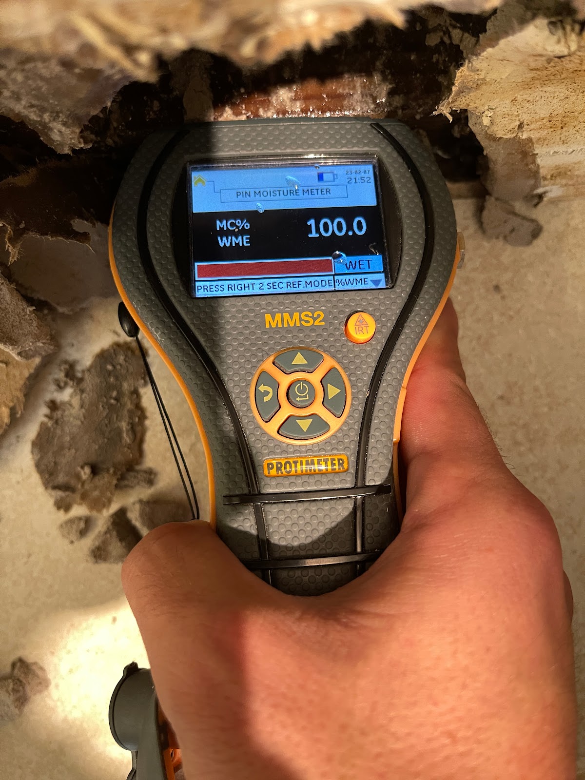 A hand holds a Protimeter MMS2 moisture meter near a wall cavity that appears to have water damage and potential mold growth. The meter's screen displays a reading of 100.0% MC% WME, indicating a very wet condition. The meter is gray and orange with a digital display and control buttons. The surrounding wall material is rough and discolored, suggesting it is saturated and possibly decaying.