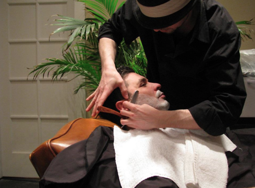 Beard grooming to the fullest; upper tapering on the cheek, and tighter tapering and hard line on top of neck. From the profile, the facial hair dips down longer around the chin, and tighter at nape of neck. A hard line (or smooth) will be made to follow easily. This includes a facial massage and hot towels.
