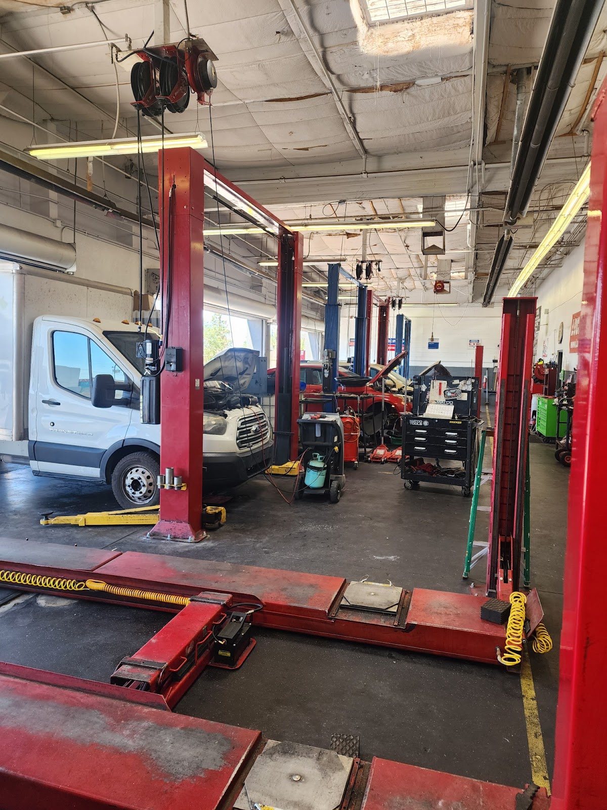 A well-lit automotive repair shop with multiple car lifts, tools, and a white van. The scene depicts a functional workspace with various equipment for vehicle maintenance.