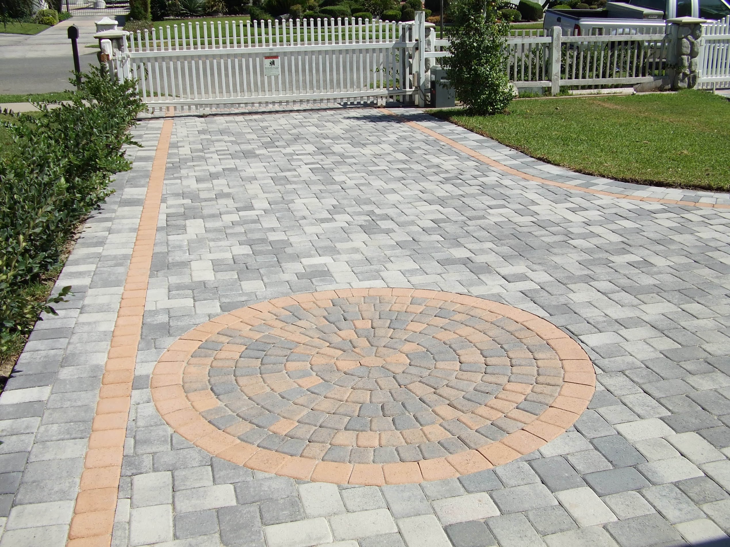 Gray paver driveway with a peach-bordered circular mosaic medallion, neat landscaping, and a white fence creating a polished residential entrance.