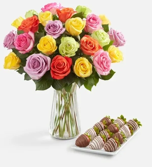 A vibrant bouquet of multi-colored roses in a clear glass vase sits next to a white platter of choco 