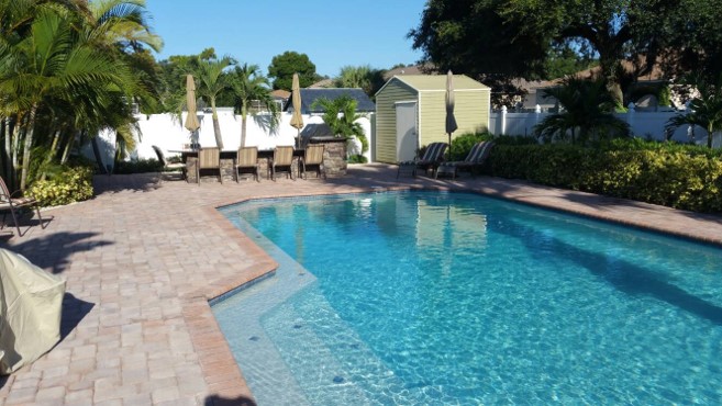 Residential backyard swimming pool with clear blue water, brick paver decking, lounge chairs, and tropical landscaping.