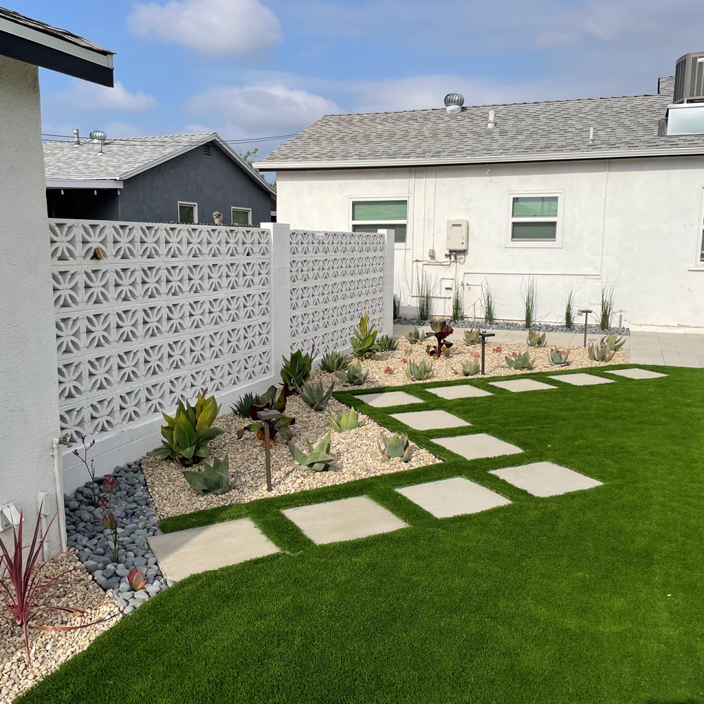Modern backyard with bright artificial turf, stepping-stone paths, drought-tolerant succulents, gravel beds, and a decorative breeze-block wall.