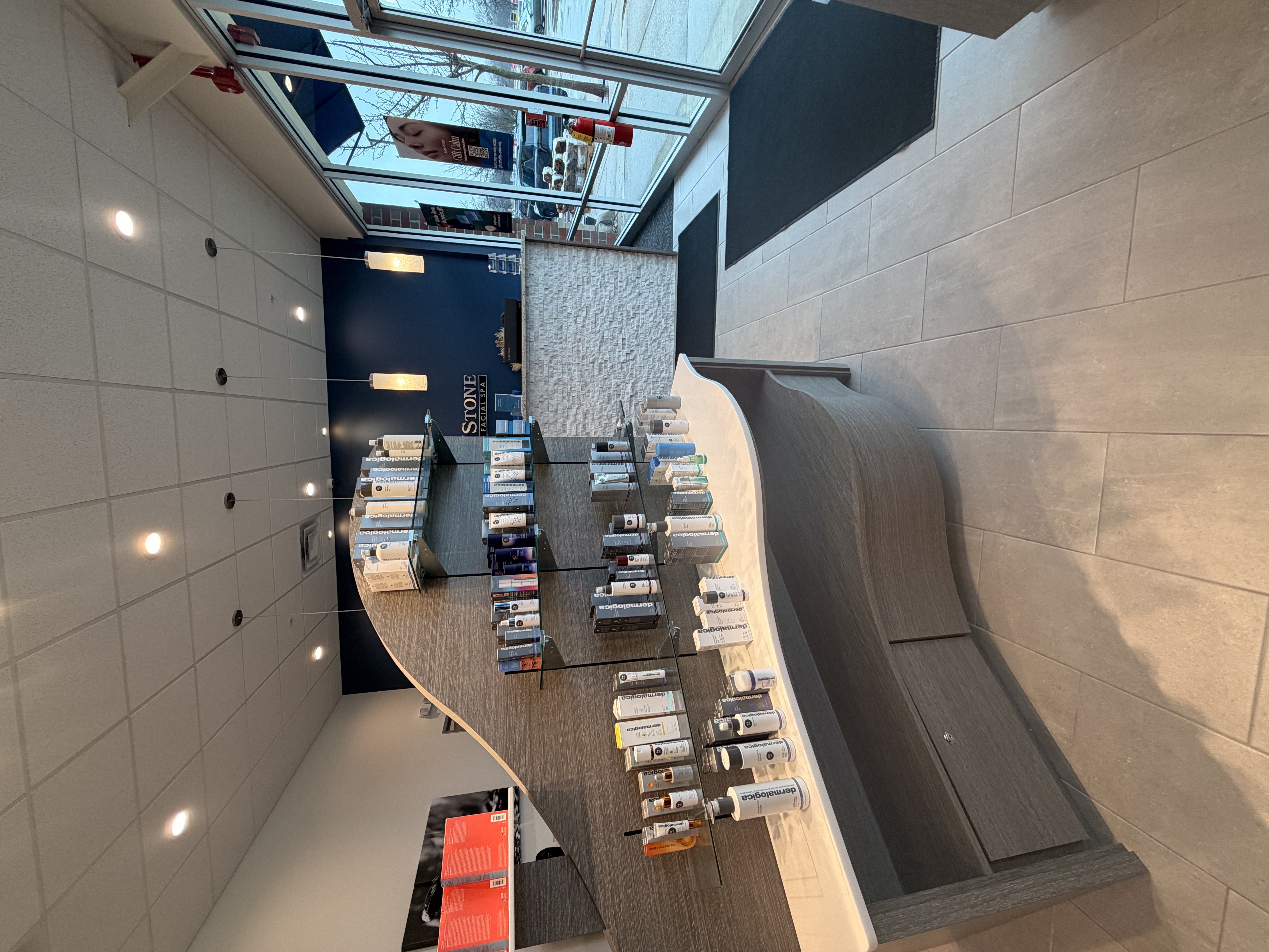 Curved retail display unit with glass shelves holding Dermalogica and skincare products. Gray wood-grain base with white countertop. Stacked stone wall and navy blue accent wall with Hand & Stone signage. Skylight windows provide natural illumination.