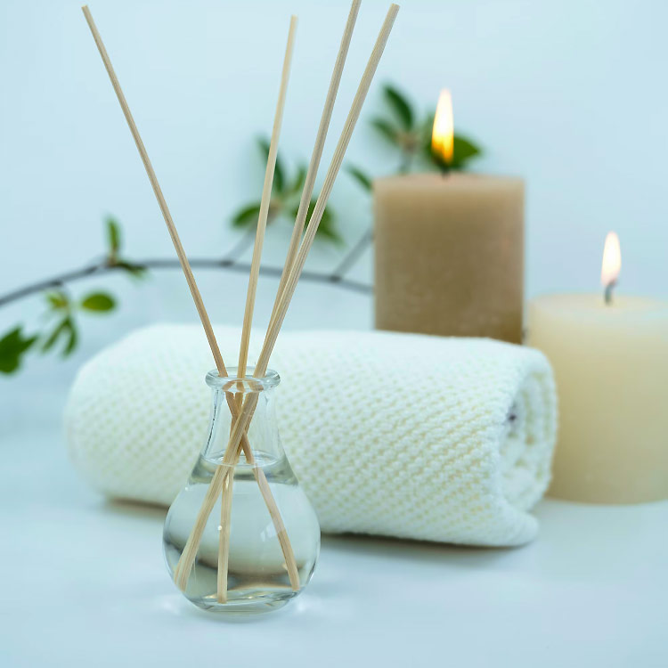 A still life arrangement for a spa or relaxation setting, featuring a glass diffuser with reeds, a neatly rolled white towel, and two lit candles in varying shades of beige and cream. A delicate branch with green leaves is visible in the background, adding a touch of nature to the serene composition.
