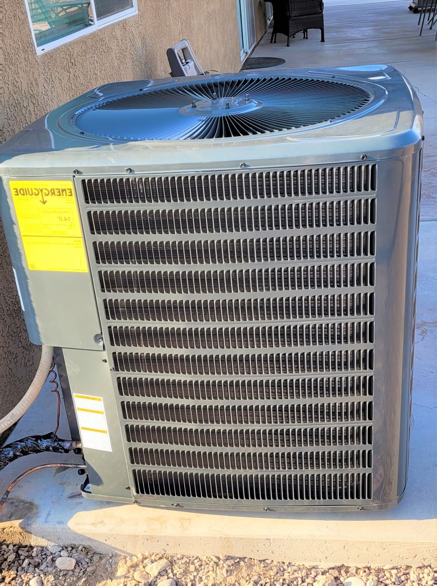 Outdoor residential air conditioning condenser unit installed on concrete pad beside home wall, featuring protective grille, top-mounted fan, and visible refrigerant lines, representing cooling system setup and HVAC installation.