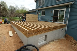 Two workers build a wooden deck frame attached to a residential home, creating the foundation for an outdoor living space. The frame sits on a concrete base, demonstrating the early stages of construction.