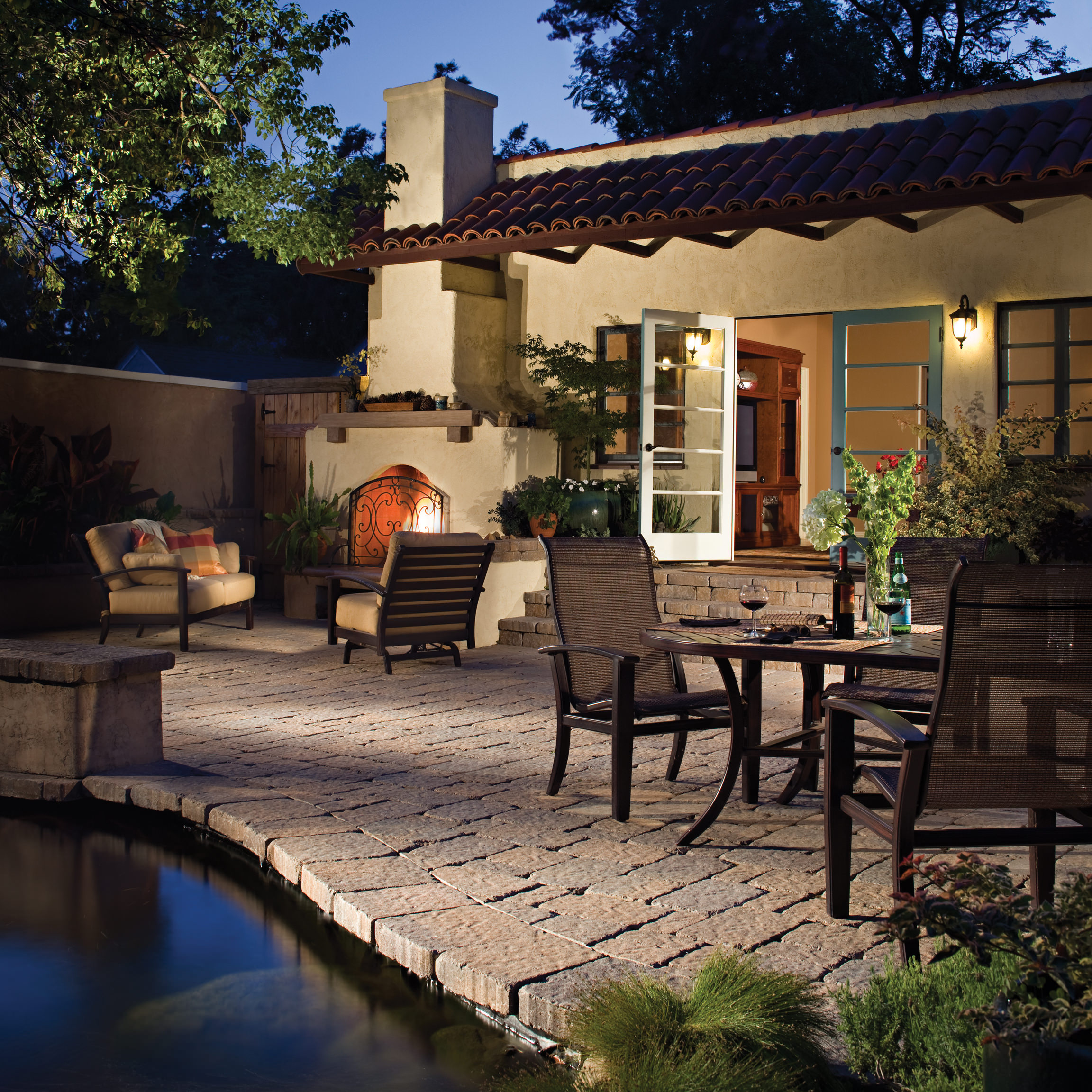 Warm, sunlit Spanish-style patio beside a pool, with terracotta roof, open glass door, fireplace, cushioned chairs, and dining set.