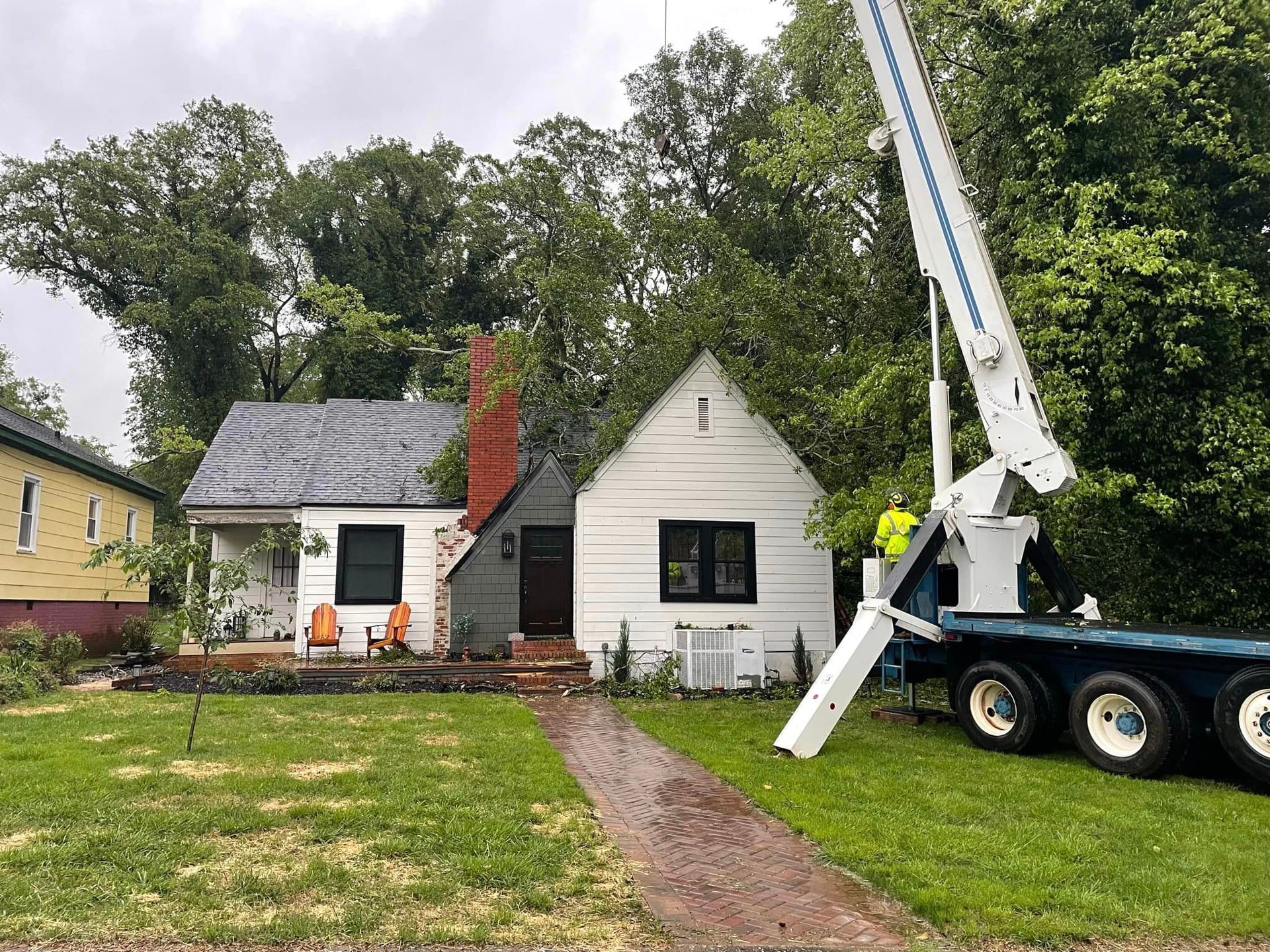 Crane-assisted tree removal taking place beside a residential house, with a worker operating equipment and large branches overhead, showing professional arborist service handling hazardous tree conditions near property.