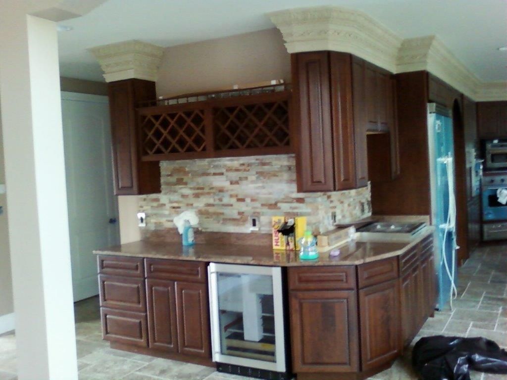 A custom kitchen setup featuring rich wooden cabinetry, stone backsplash, and built-in wine rack. The island includes storage and a mini fridge, combining functionality with elegant craftsmanship and modern design elements.