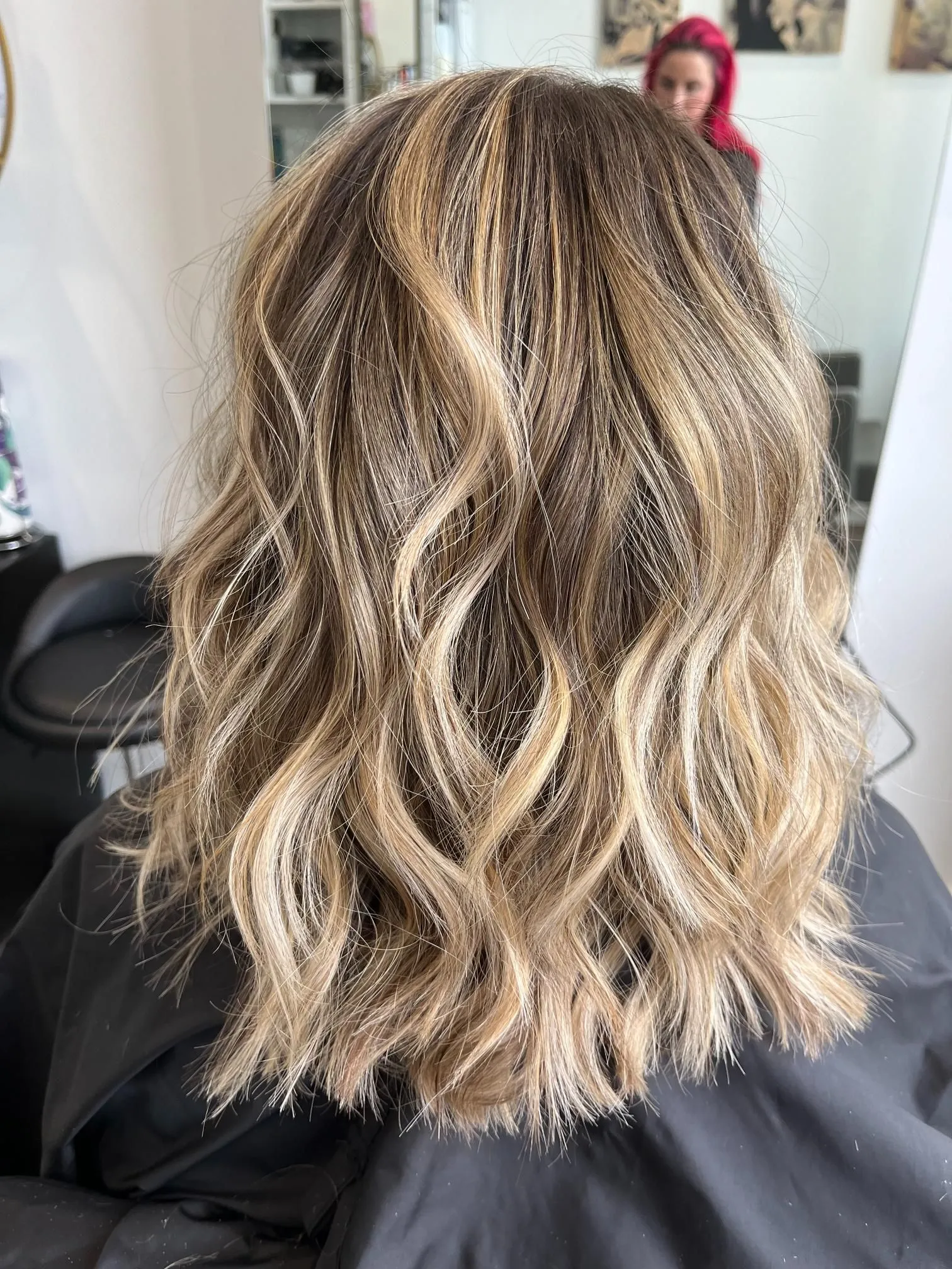 Freshly styled shoulder-length blonde balayage hair with soft waves, showcasing natural dimension and highlights. The hairstyle is photographed from the back inside a salon, highlighting professional coloring and styling results.
