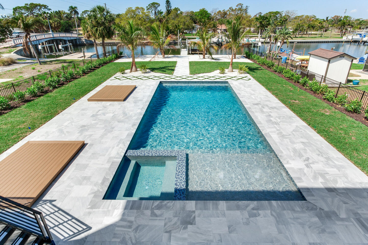 Luxury backyard pool and spa area with a spacious paver patio, manicured lawn, tropical landscaping, and waterfront views.