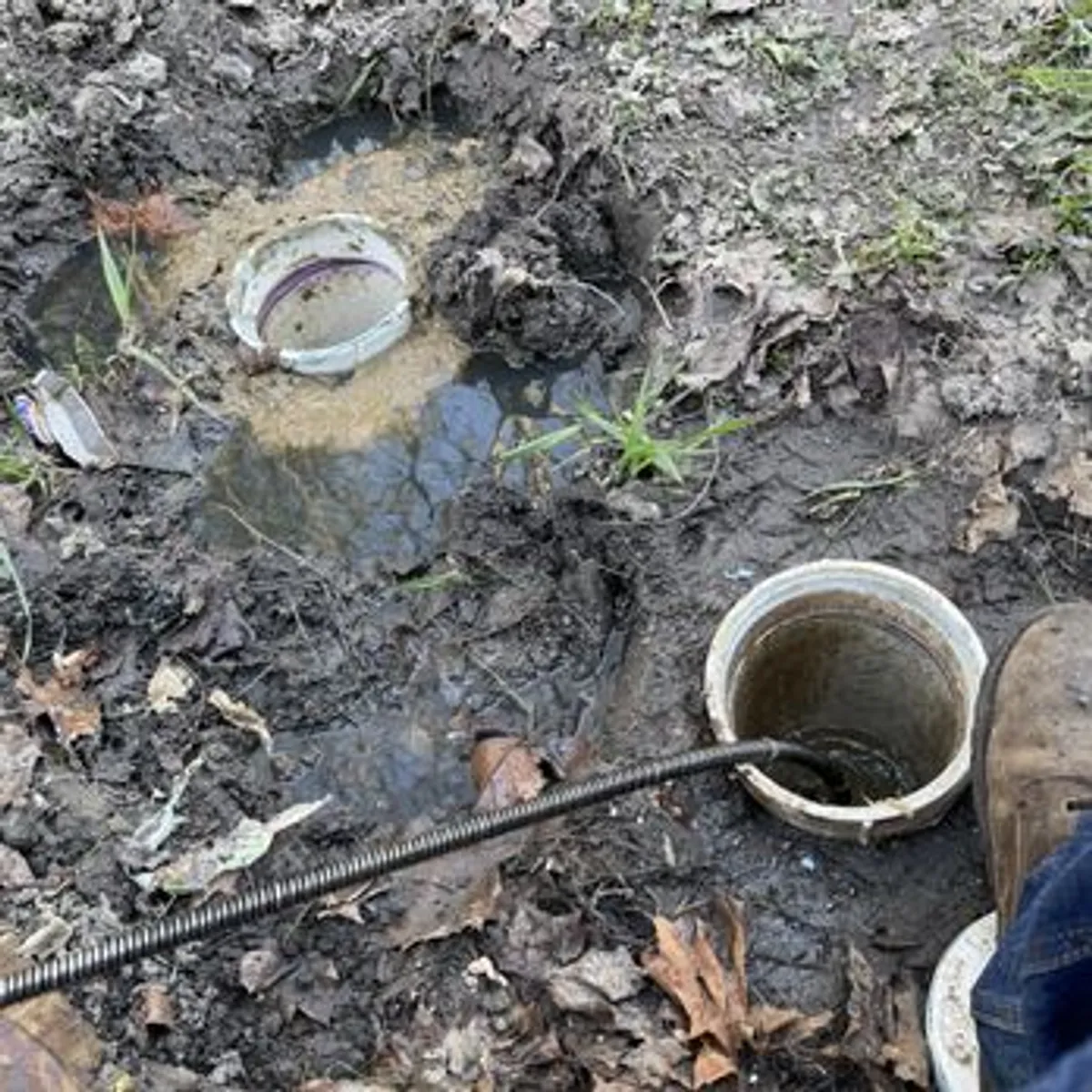 A drain snake is inserted into a pipe opening in muddy ground, with another pipe opening visible nearby. The scene appears to be outdoors in a messy, wet environment.
