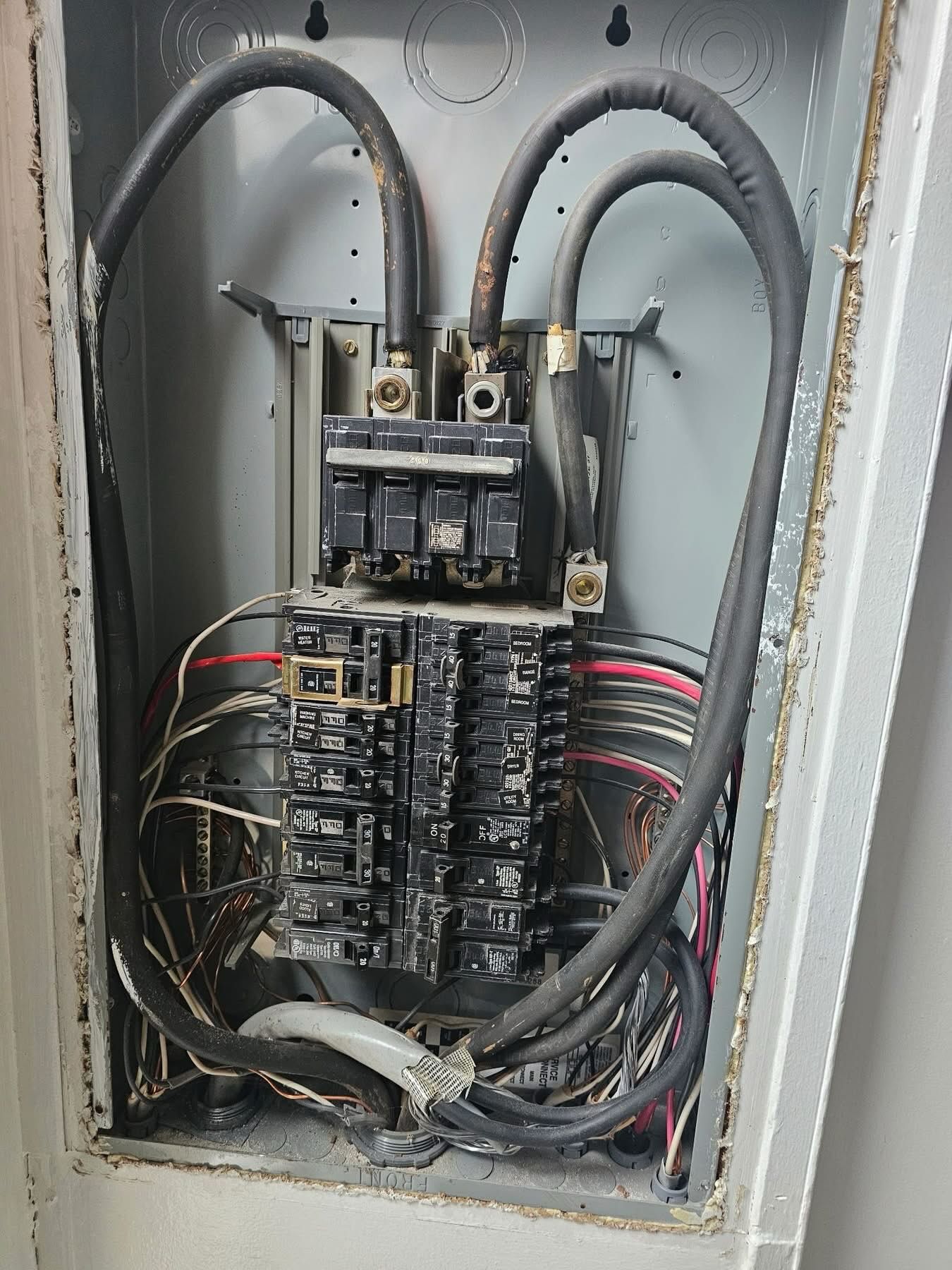 Open electrical breaker panel showing multiple circuit breakers, thick service cables, and wiring connections, illustrating interior layout of residential electrical system with organized but aged components and visible cable routing.