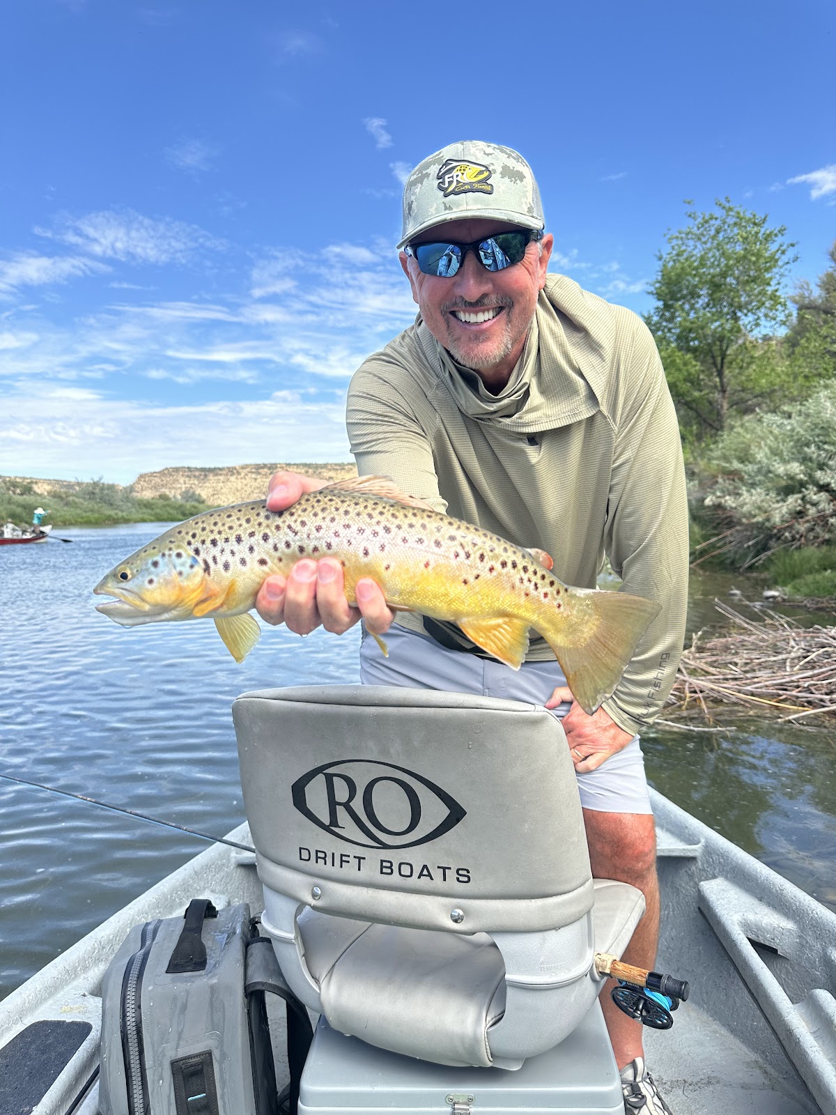 A man in a camouflage hat and sunglasses smiles while holding a large trout he just caught while fishing from a boat on a lake under a blue sky with fluffy white clouds. He is wearing a light-colored fishing shirt and shorts. The boat has a seat with a logo that reads "RO DRIFT BOATS" and a fishing rod is visible in a holder on the side of the boat.
