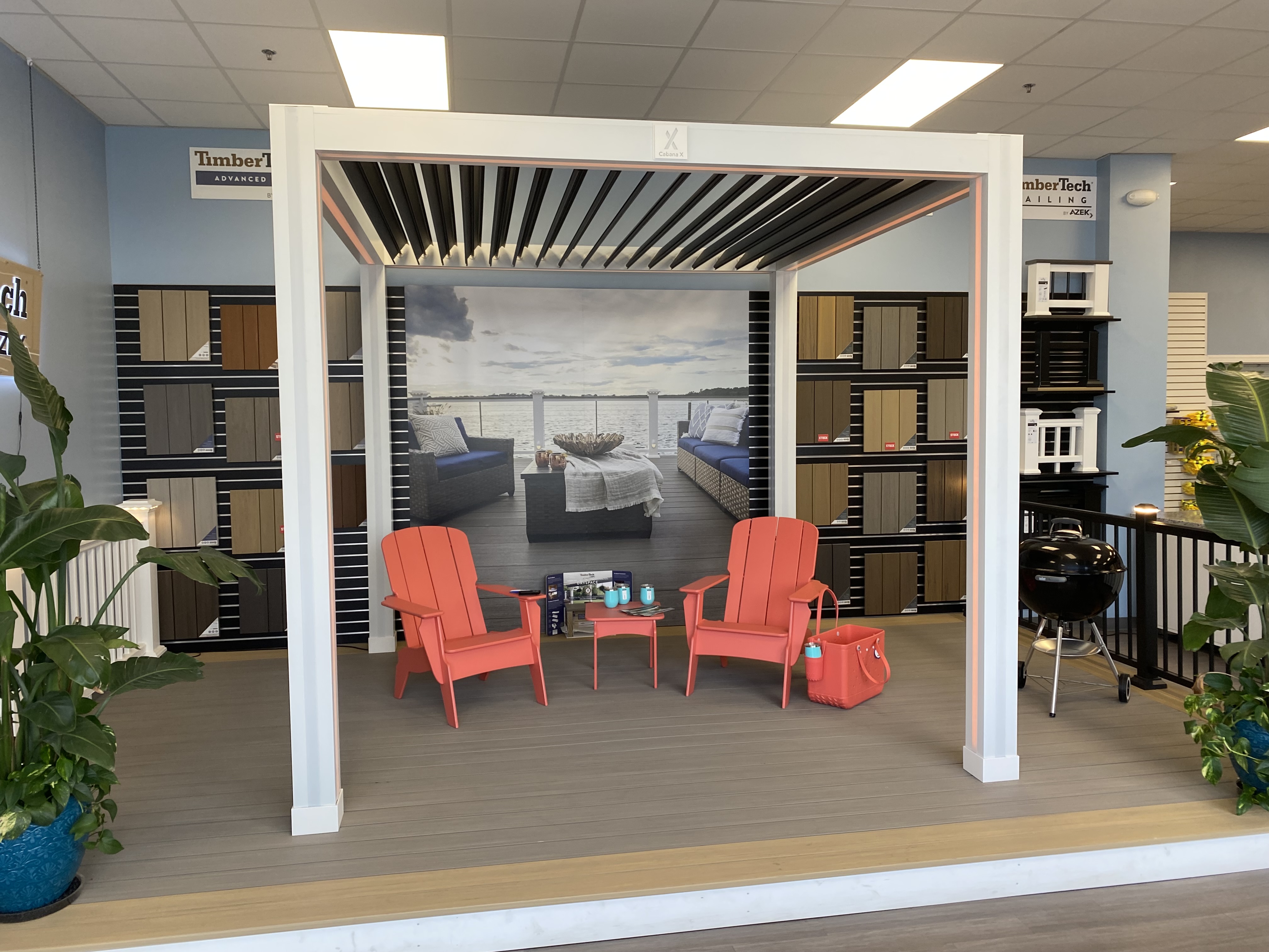 A showroom display featuring a stylish outdoor living setup with composite decking, a modern pergola, and vibrant chairs. The backdrop showcases a scenic waterfront view, while wall-mounted decking samples highlight available colors and materials. Ideal for customers exploring premium decking and millwork solutions.