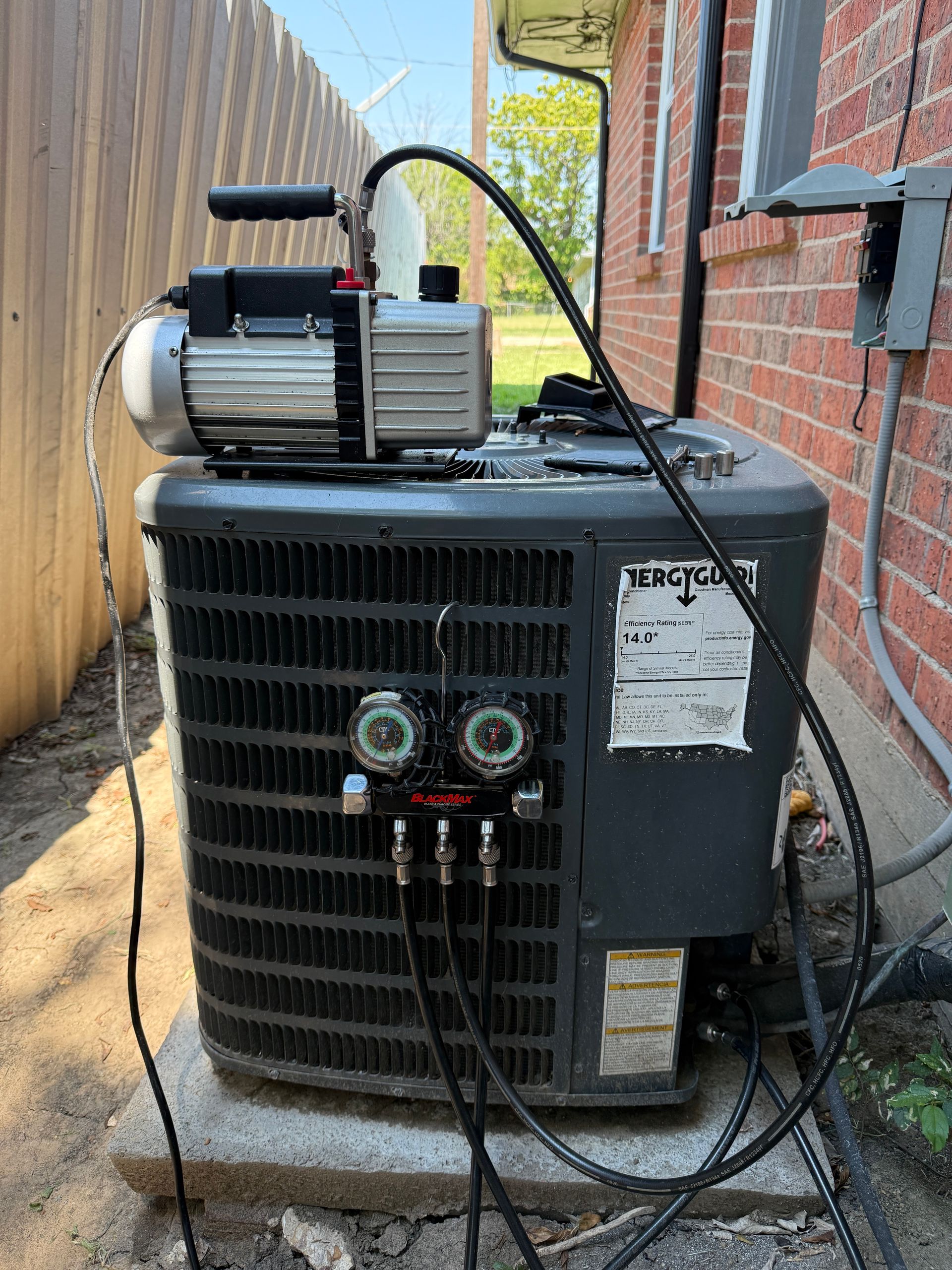 Outdoor condenser unit connected to gauges and vacuum pump for refrigerant servicing. Technician equipment indicates system charging, pressure testing, or evacuation during professional HVAC maintenance near residential building.