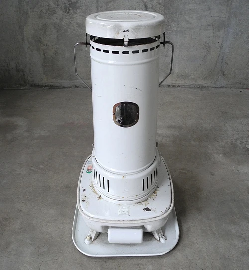 A vintage, white kerosene heater stands on a concrete floor, showcasing a simple yet practical design. The cylindrical heater features a small window for viewing the flame, and it sits atop a sturdy base with handles on either side for portability.