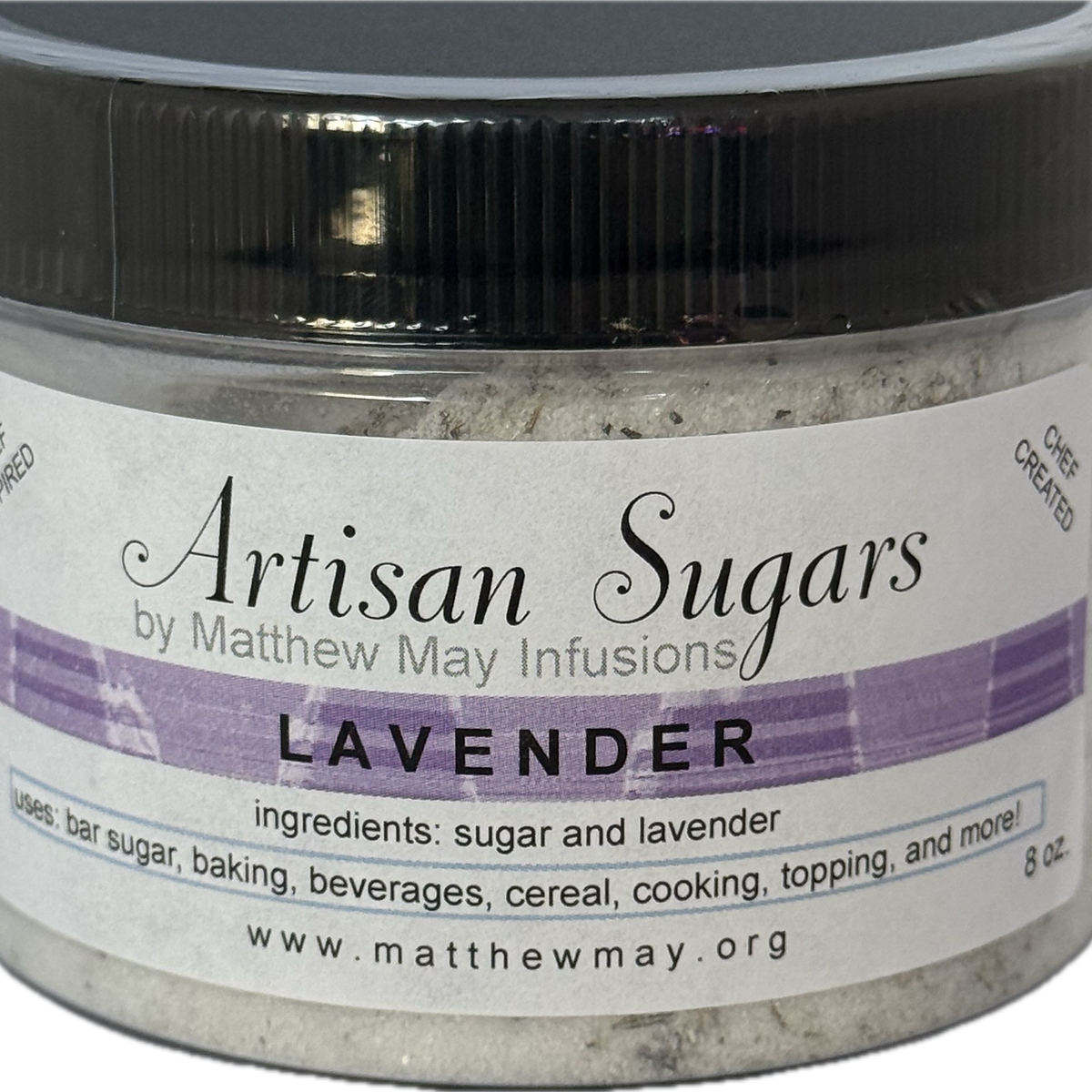 A close-up shot of a jar of Artisan Sugars, specifically the Lavender flavor, made by Matthew May Infusions. The jar has a black lid and a white label with a purple striped band. The label details the ingredients as sugar and lavender, and lists various uses such as in bar sugar, baking, beverages, cereal, and as a topping. The product is described as 'CHEF CREATED' and the website 'www.matthewm a y.org' is visible.