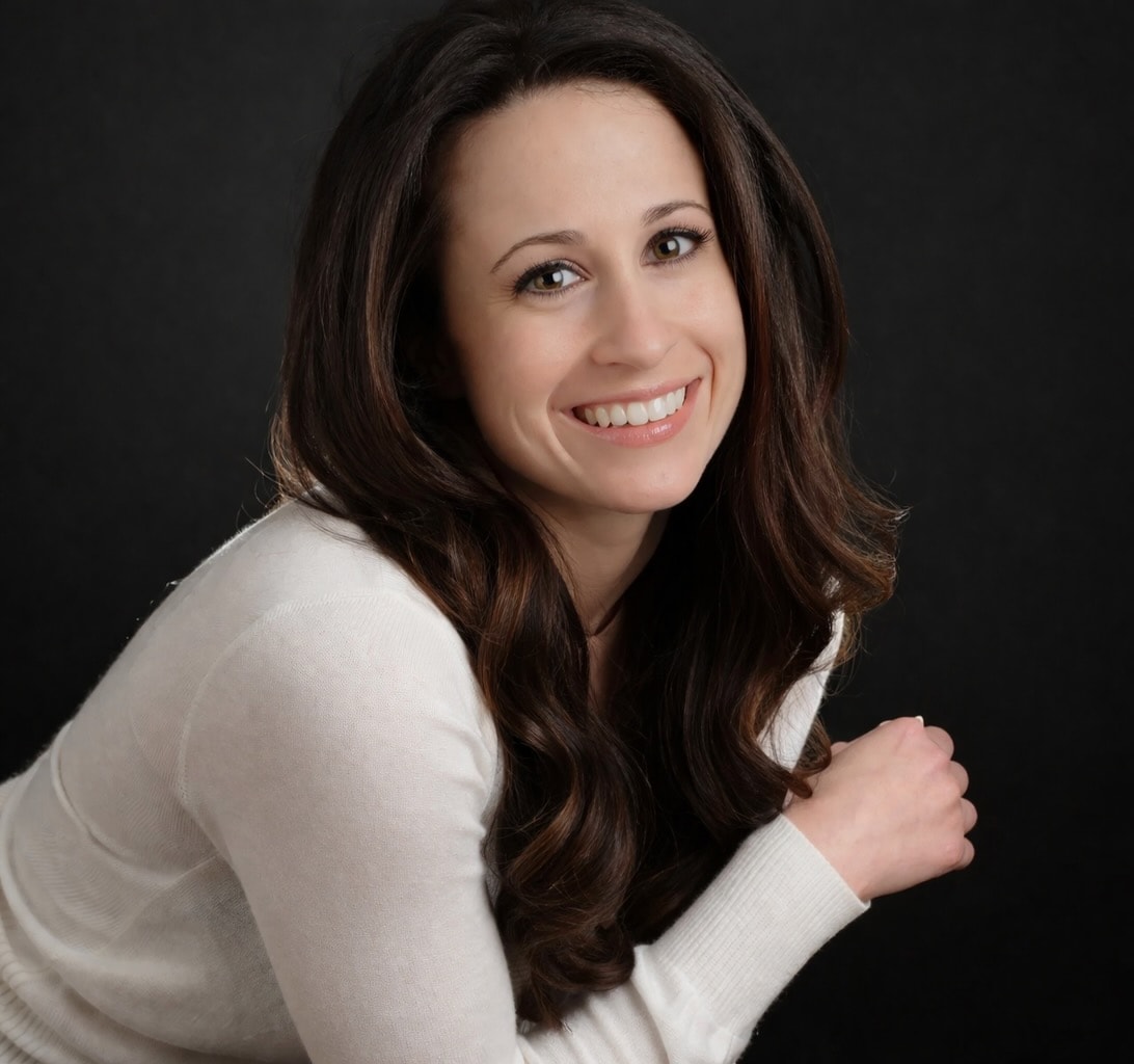 A medium shot portrait of a young woman with long, wavy brown hair and brown eyes. She is smiling broadly and looking directly at the camera. She is wearing a cream-colored sweater with a crew neck. The background is a dark, solid color, likely black or dark gray.