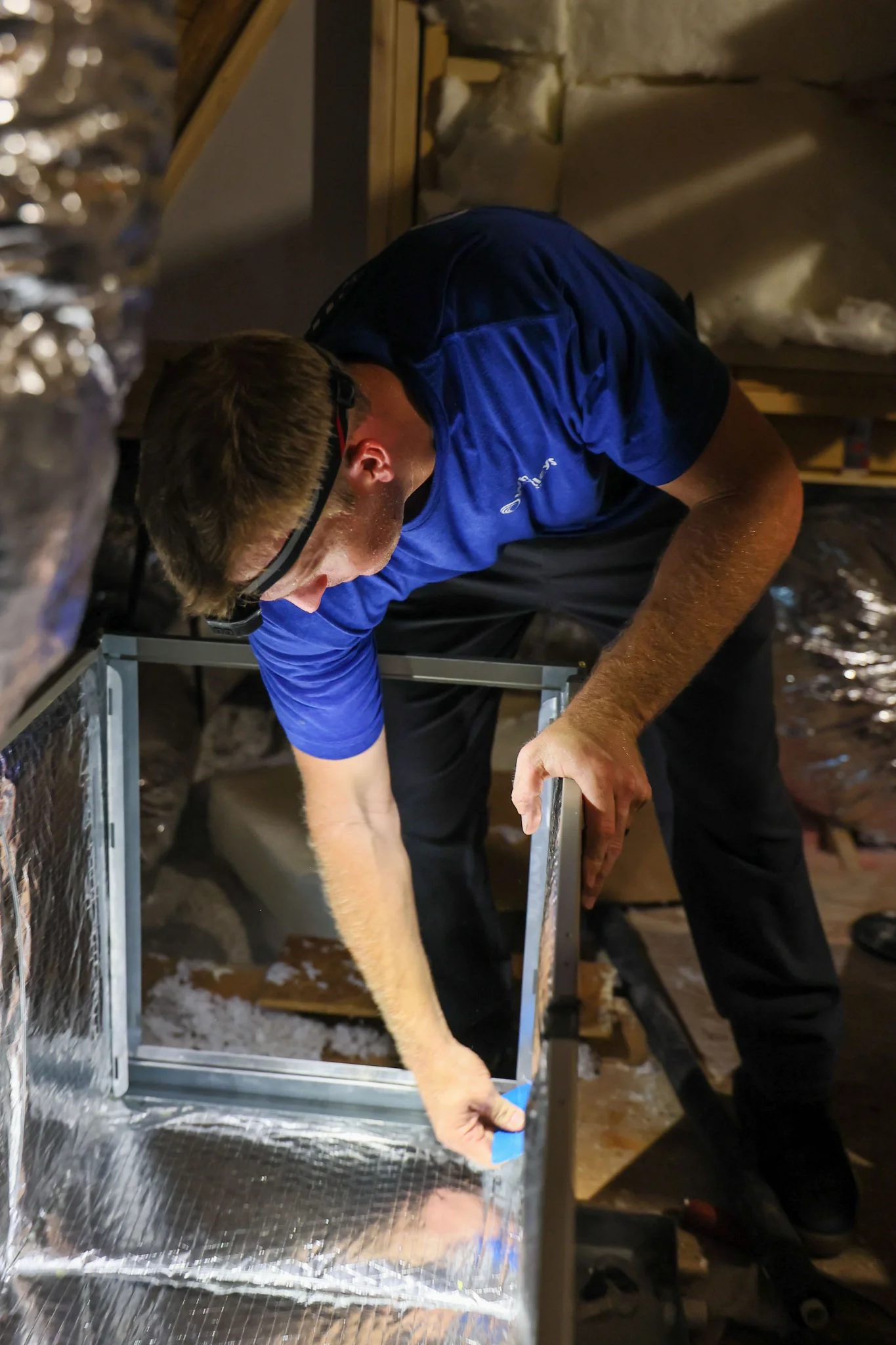 A man wearing a blue shirt and dark pants, with a headlamp on, is working in an attic. He is bending 