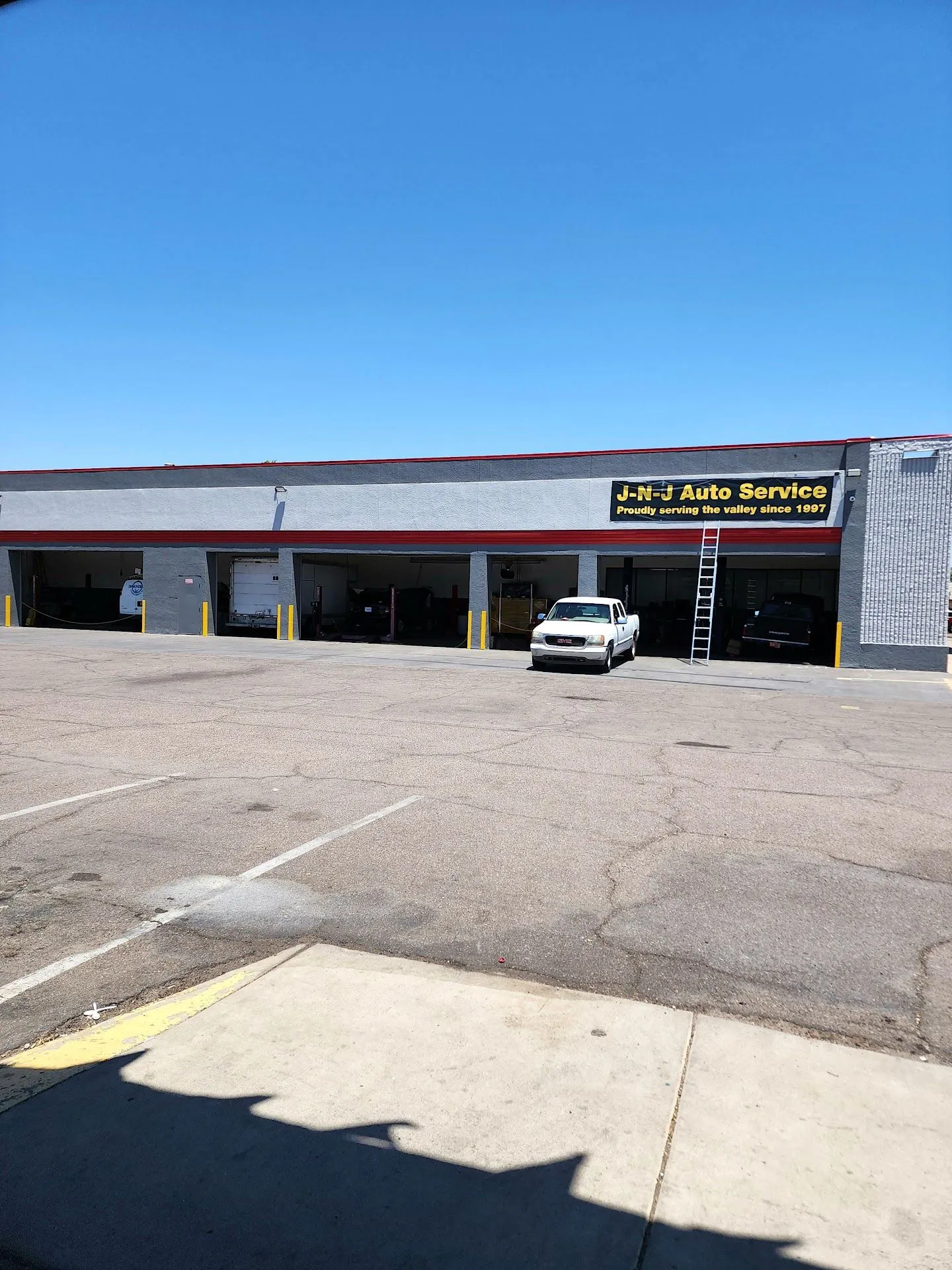 A white GMC pickup truck is parked in front of J-N-J Auto Service, a business with multiple garage bays. A tall ladder is propped against the building near the truck. The building has a gray exterior with a red trim line and a yellow banner that reads "J-N-J Auto Service Proudly serving the valley since 1997". The sky is clear and blue, and the ground in front of the building is a cracked asphalt parking lot. The image appears to be taken on a sunny day.