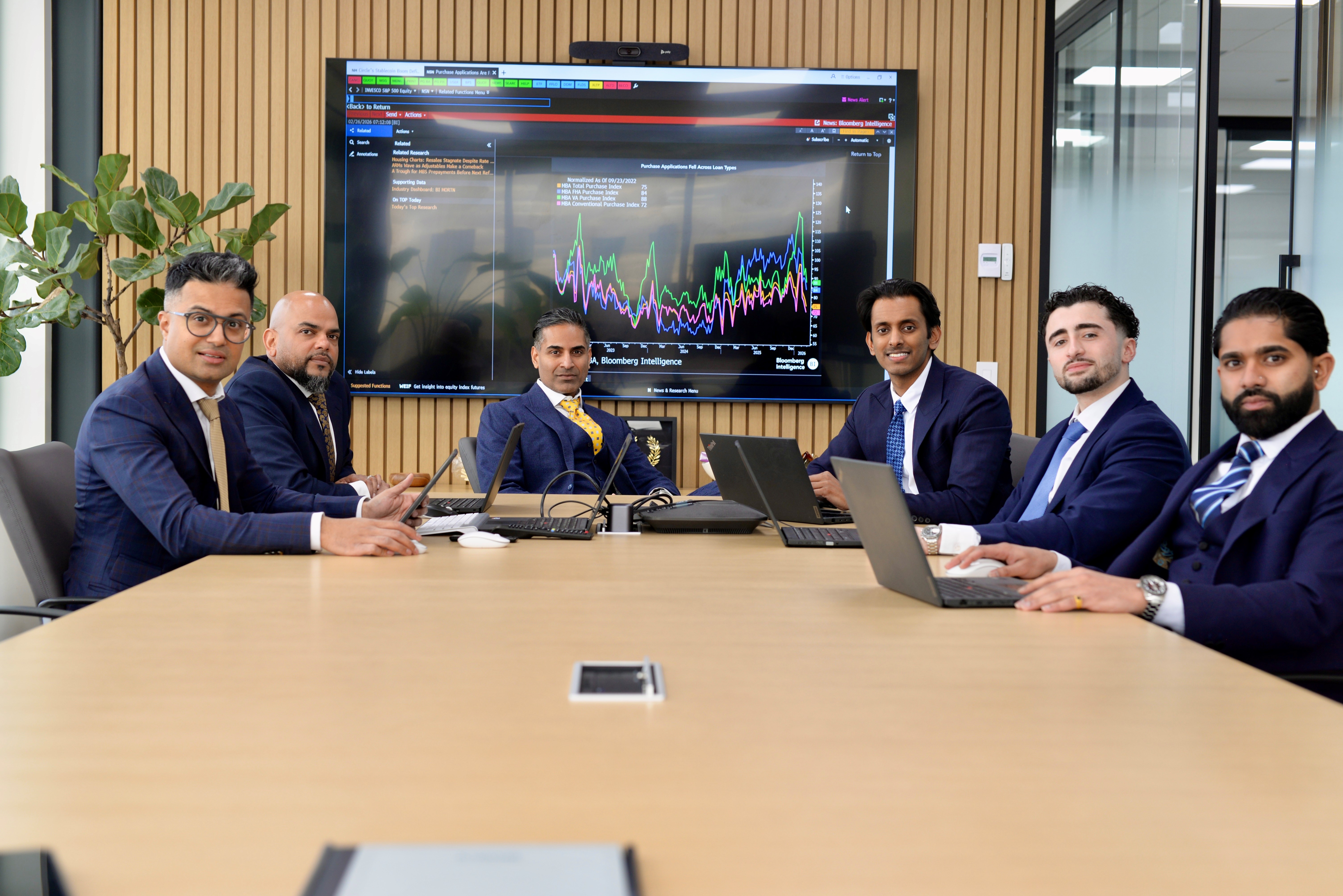 Six financial advisors from the Ilangovan Wealth Group in a modern conference room, seated around a table with laptops, reviewing Bloomberg Intelligence market data displayed on a large wall-mounted screen. The screen shows multiple colorful trend lines tracking financial performance metrics. The contemporary office features a wooden slat accent wall and natural light from glass walls.