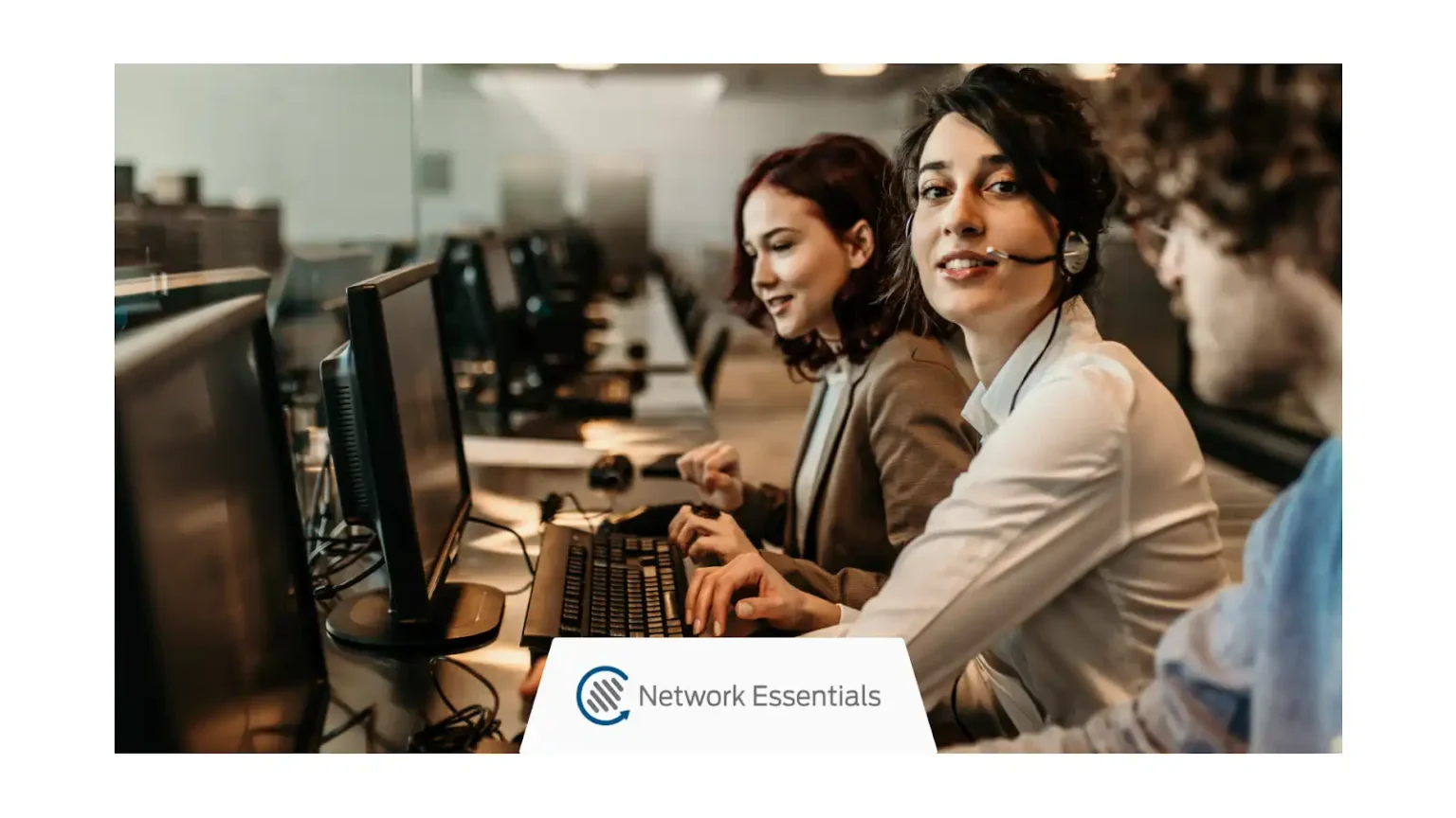 A diverse group of three customer service representatives are working at computers in a brightly lit office. The woman in the foreground on the right, wearing a headset, smiles at the camera while looking slightly to the left. To her left, another woman in a brown blazer is focused on her computer screen, typing on the keyboard. A third person, partially visible on the right, is also looking at a screen. The office environment appears to be a call center with rows of computers. A logo for "Network Essentials" is visible in the lower center of the image.