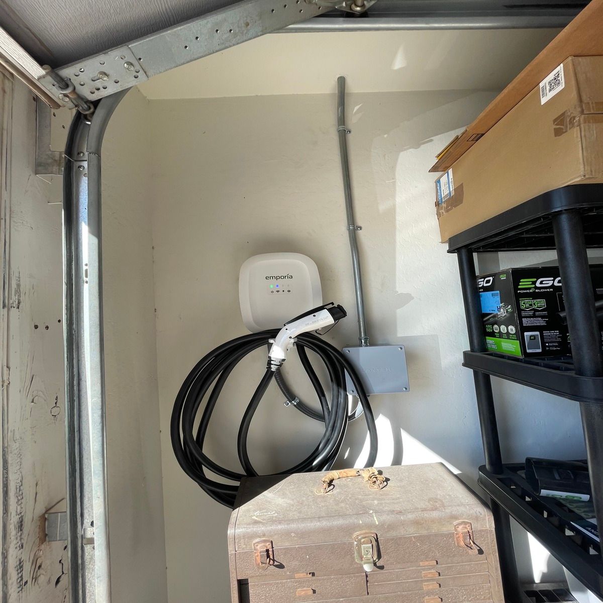 An Emporia electric vehicle charger is installed inside a garage, with black charging cable looped around the wall-mounted unit beside electrical conduit, junction box, shelving, tools, and storage equipment nearby.