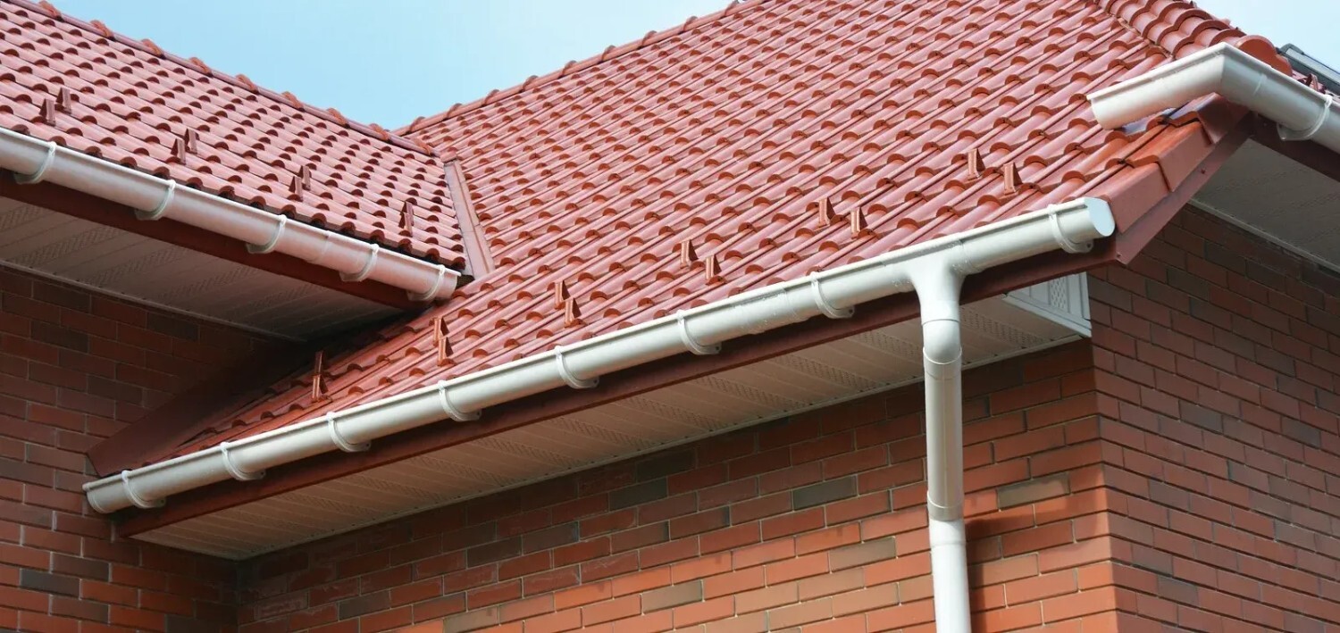 A house with a red tiled roof, beige gutters, and downspouts, featuring snow guards for safety and a brick exterior.