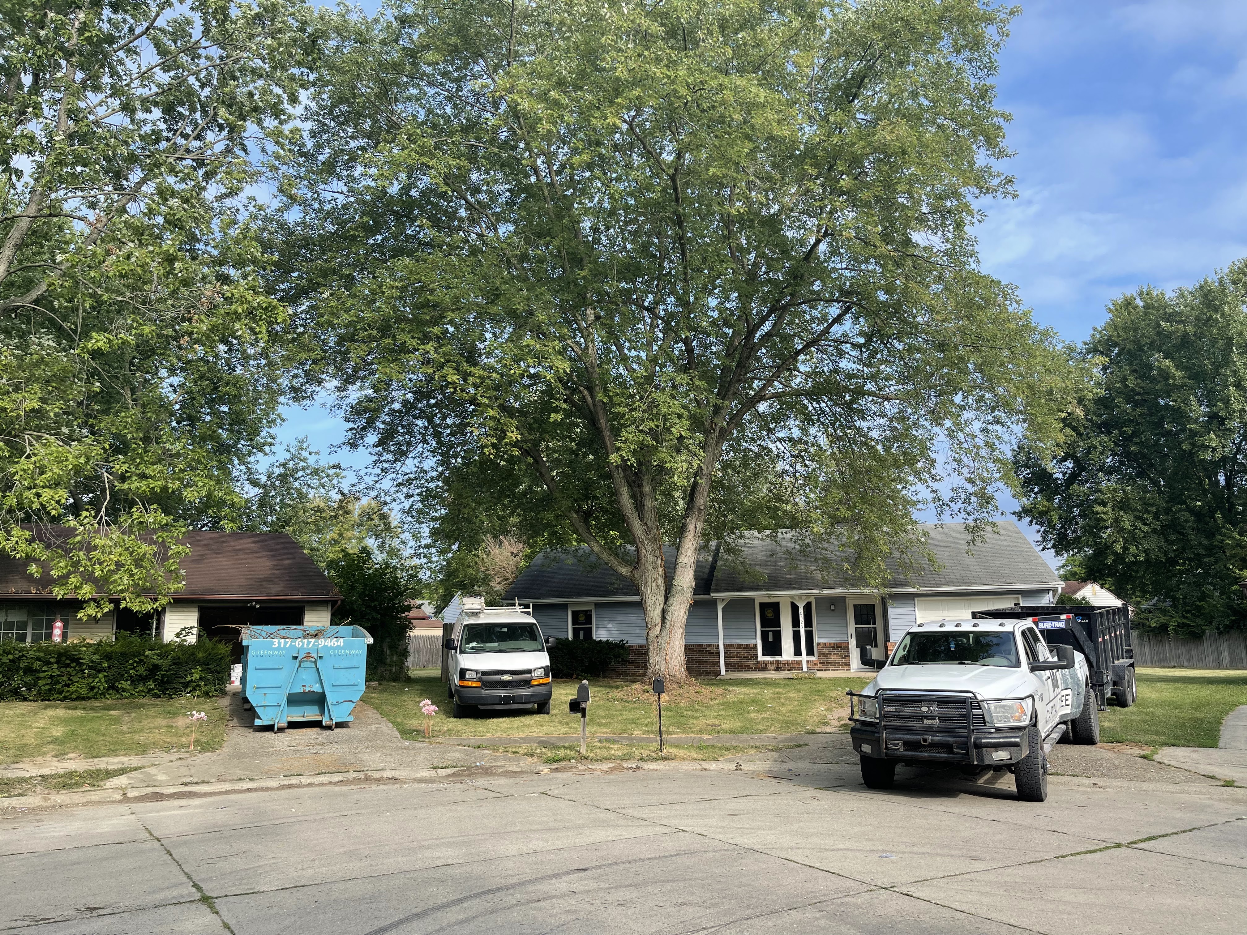 A residential street scene with two houses, several vehicles including a white pickup truck with a trailer and a white van, and a large blue dumpster. A large tree dominates the center of the frame, with its branches extending over the houses and street. The sky is blue with some scattered clouds.