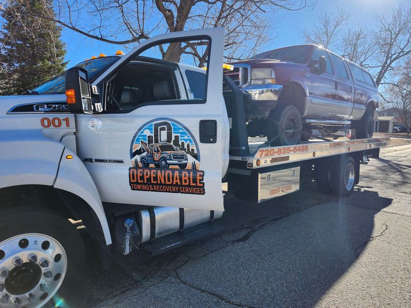 Dependable Towing & Recovery Services truck carries a damaged pickup on a flatbed during daytime, highlighting professional vehicle recovery and transport services in a residential neighborhood setting.