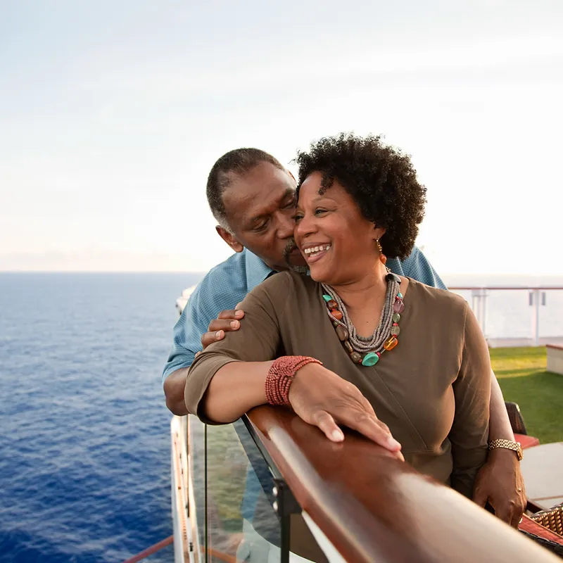 A man is embracing a woman from behind as they stand on the deck of a cruise ship, with the ocean and sky in the background. The woman is laughing and looking to her right, while the man whispers something in her ear. They are both dressed casually, and the woman is wearing jewelry.