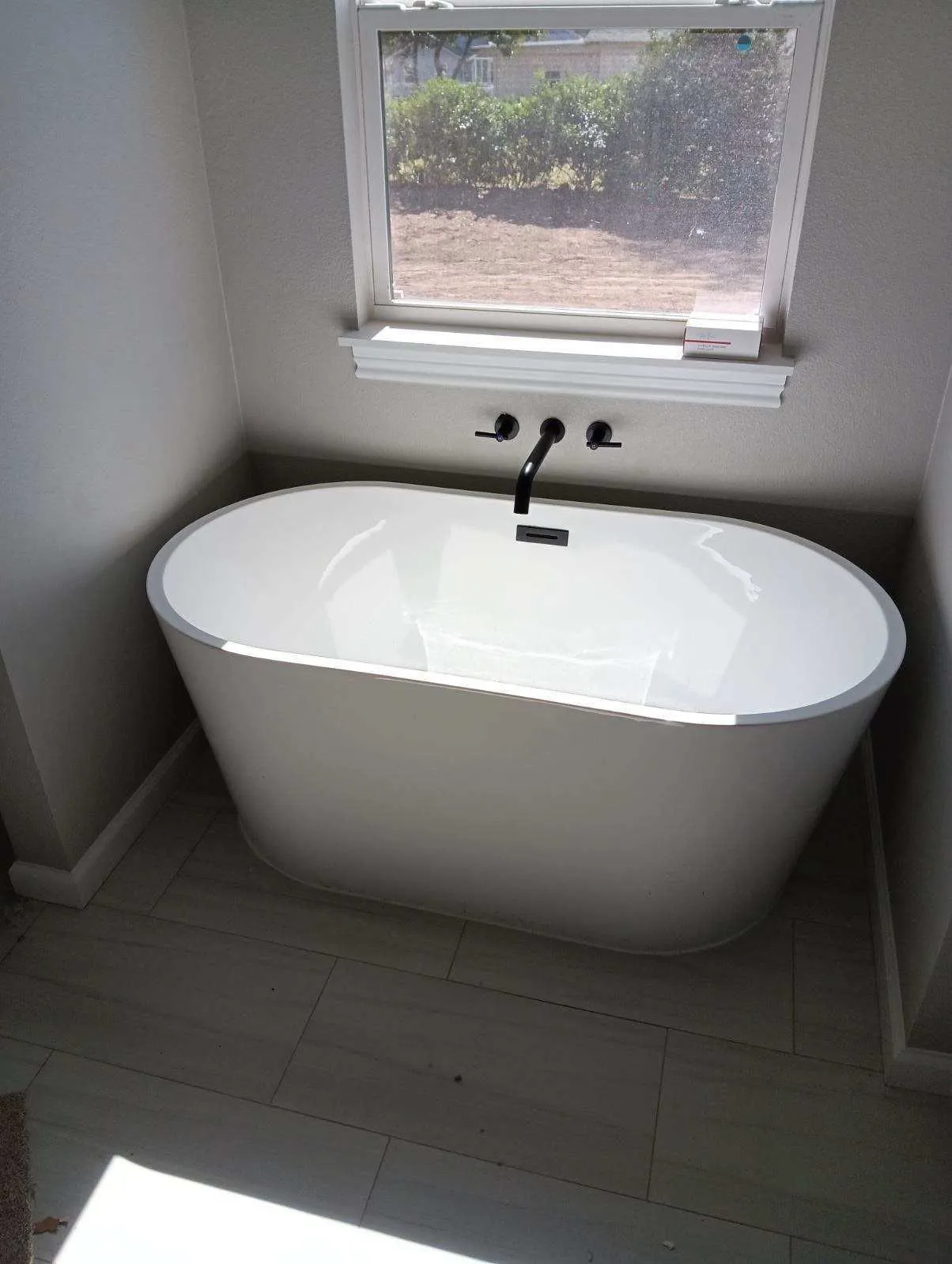 Modern freestanding soaking bathtub installed beneath large window with matte black wall-mounted faucet, creating luxurious bathroom design featuring premium plumbing fixtures, elegant aesthetics, and relaxing residential comfort for homeowners.