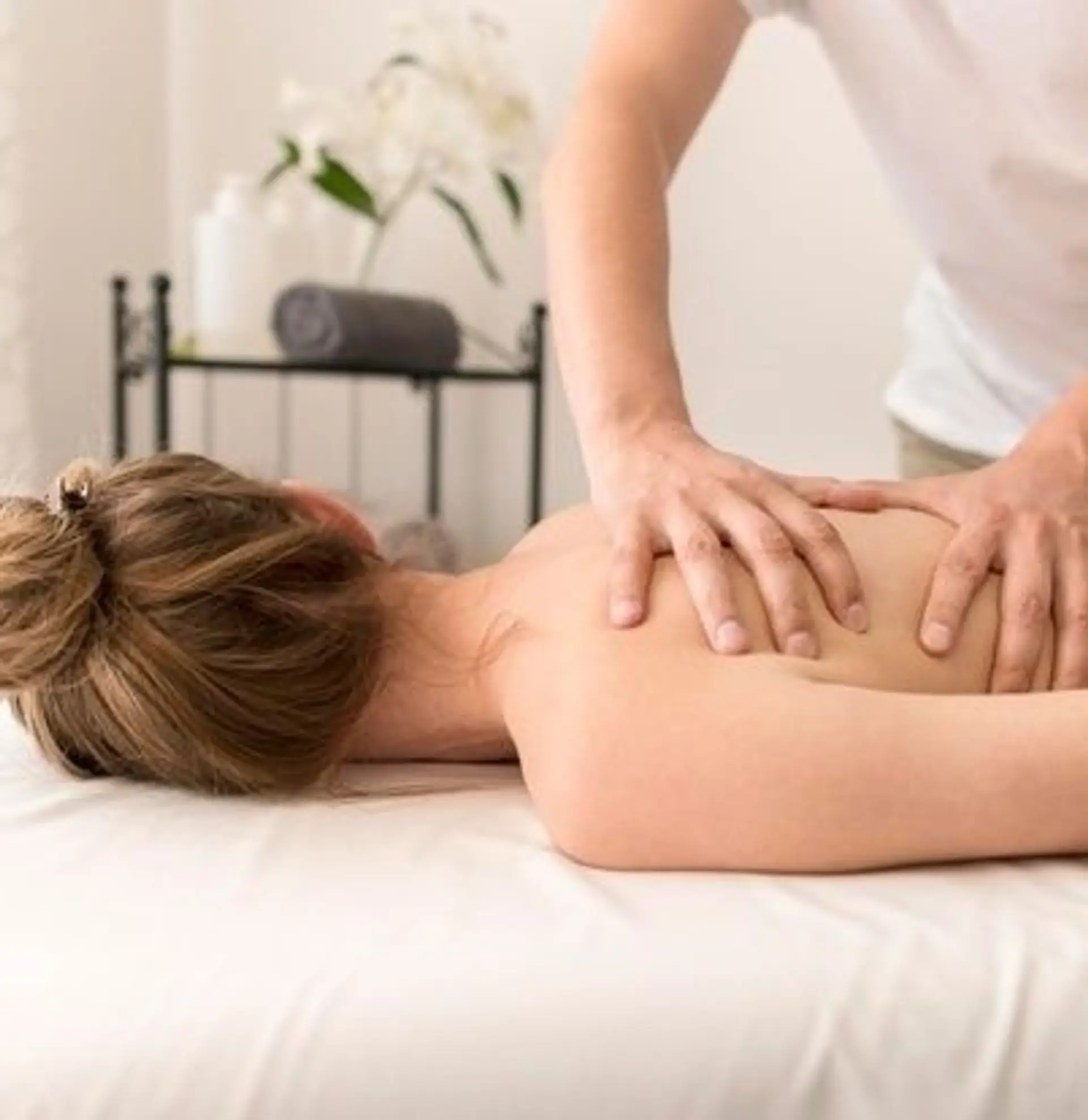 A close-up shot of a person receiving a massage on their back and shoulders. The masseuse's hands are gently applying pressure to the client's upper back, while the client is lying face down on a white massage table. In the background, a shelf holds rolled towels and white bottles, suggesting a spa or wellness setting.