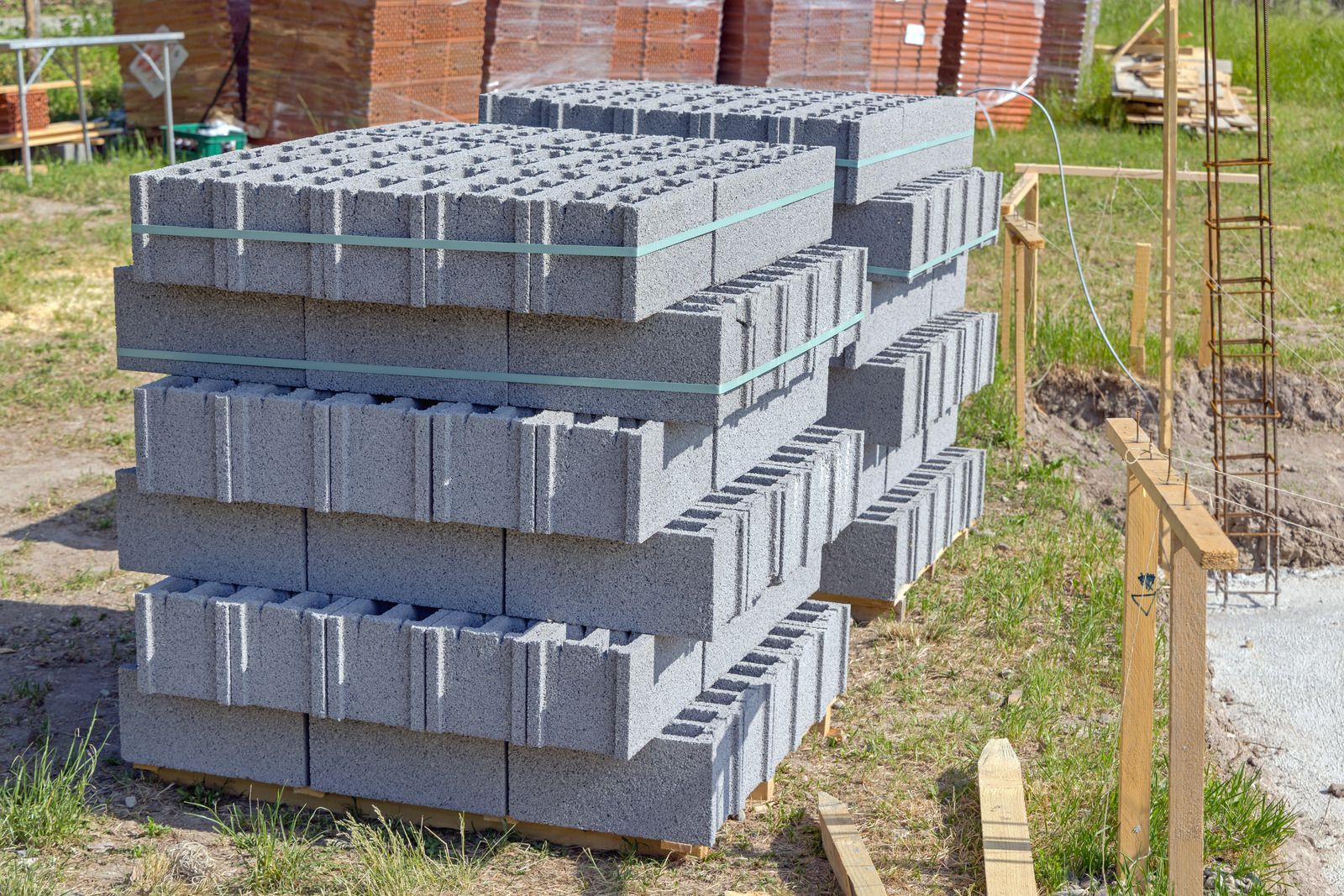 Stacked gray concrete masonry units sit on wooden pallets at a sunny construction site. The textured blocks are secured with green straps, positioned near foundation rebar and orange bricks.
