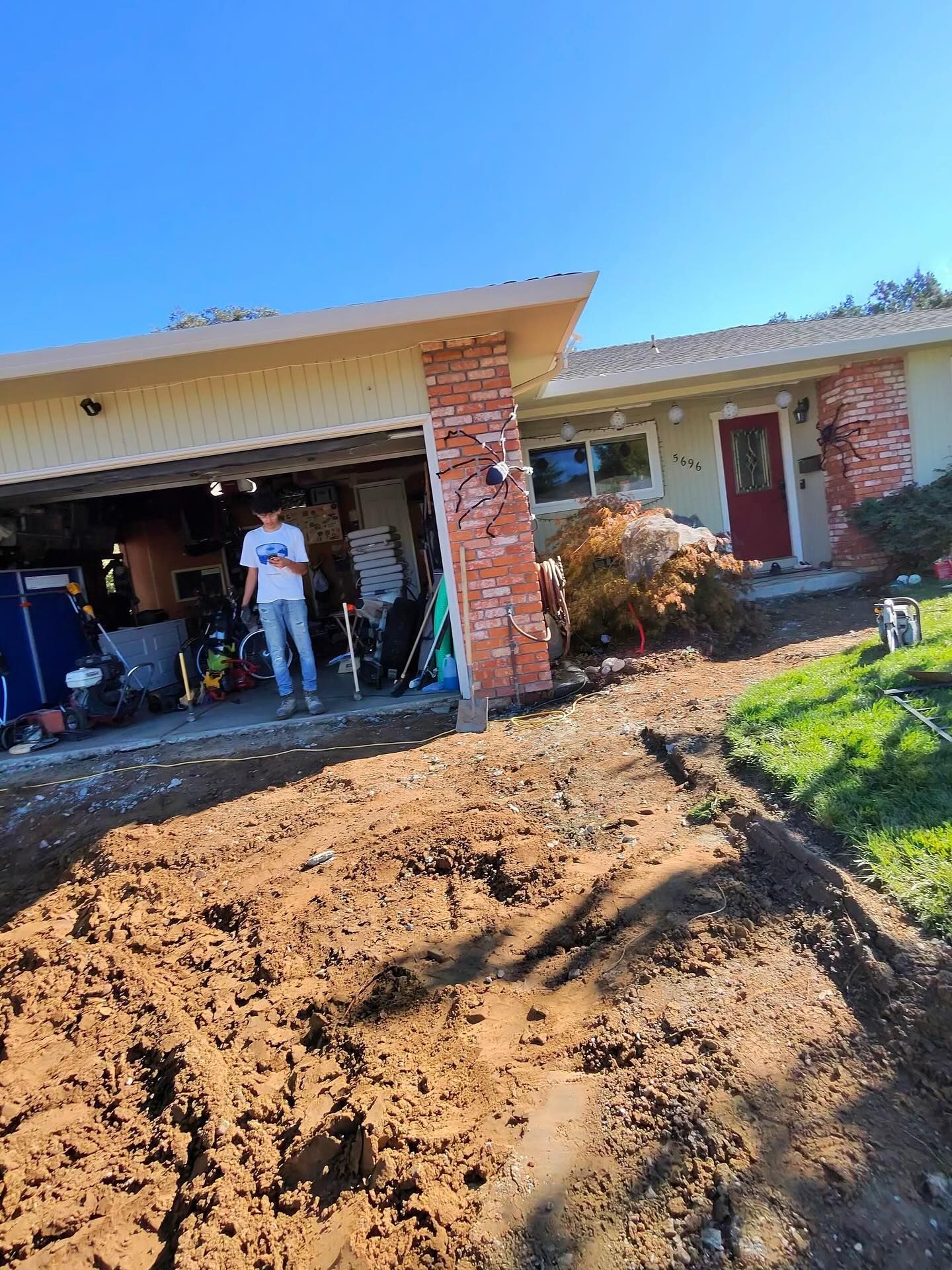 Residential front yard excavation with soil removed and driveway area cleared, showing ongoing construction work, exposed ground, and homeowner standing near open garage, highlighting site preparation and landscaping or paving project.